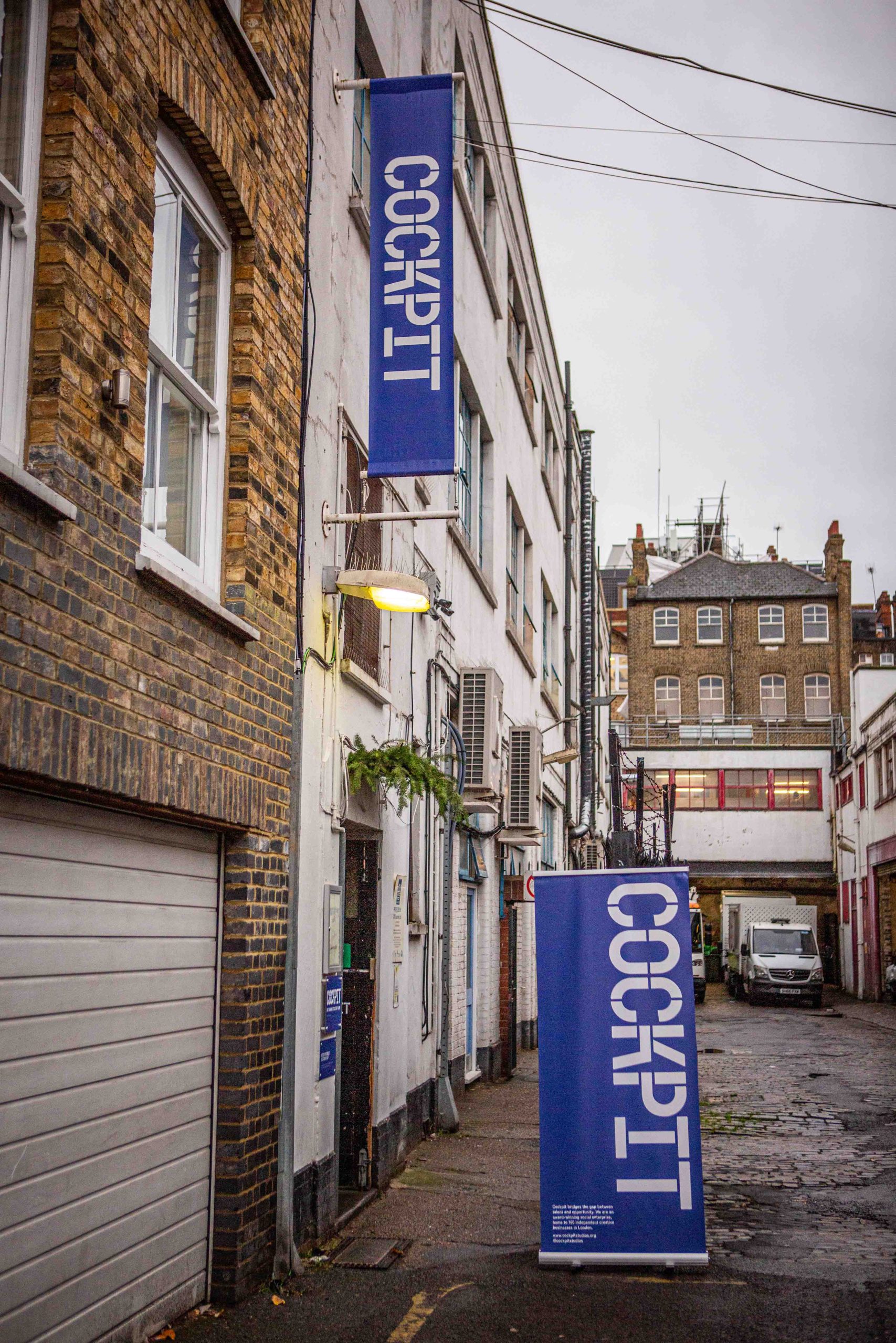 Street in Central London with brick and white buildings, featuring blue vertical signs reading 'COCKPIT' and a parked white van in the background.