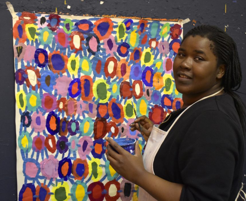 Photograph of Kwaga Sillingi looking at the camera whilst holding a paint brush and paint pot. She is stood in front of her painting.