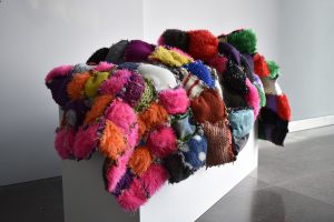 Small squares of colourful fabric sewn together to create a large artwork that is draped over a white plinth