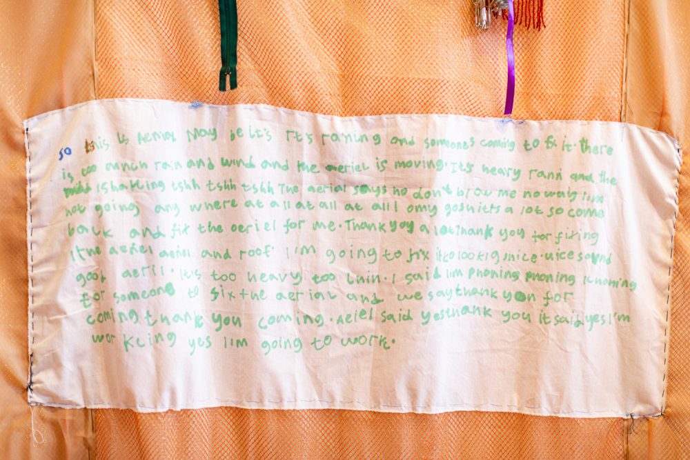 Fabric wall hanging by Andrew Omoding. Text is wirtten on the central panel of the work