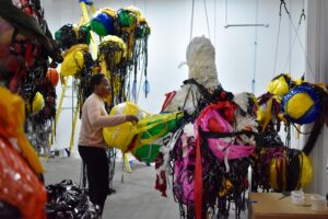 Nnena Kalu standing and building a boulder onto her suspended artworks make out of layers of tape, paper and plastic.