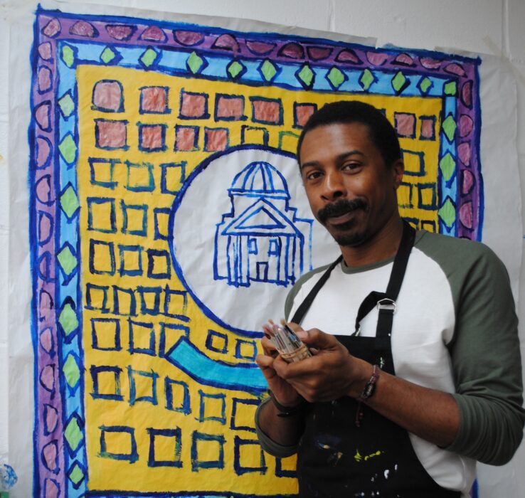 Thompson Hall standing in front of his painting. Thompson is wearing a black apron and is holding a selection of small paint brushes 
