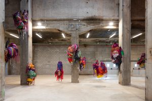 Several colourful mixed media sculptures suspend from the ceiling in a large industrial hall.