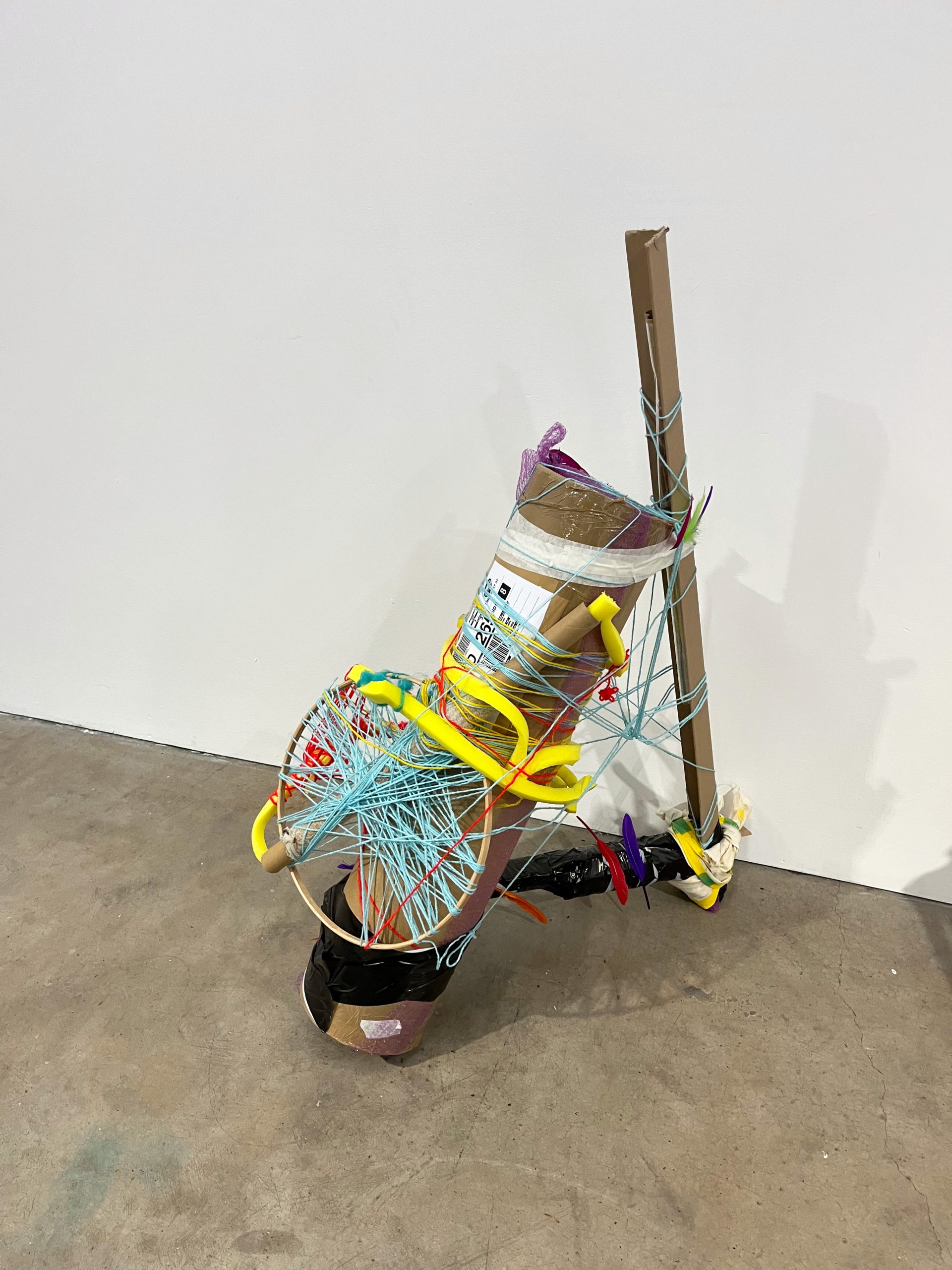An abstract sculpture rests against a white wall on a concrete grey floor. The sculpture is assembled of various materials i.e. brown cardboard, black tape, blue string. 