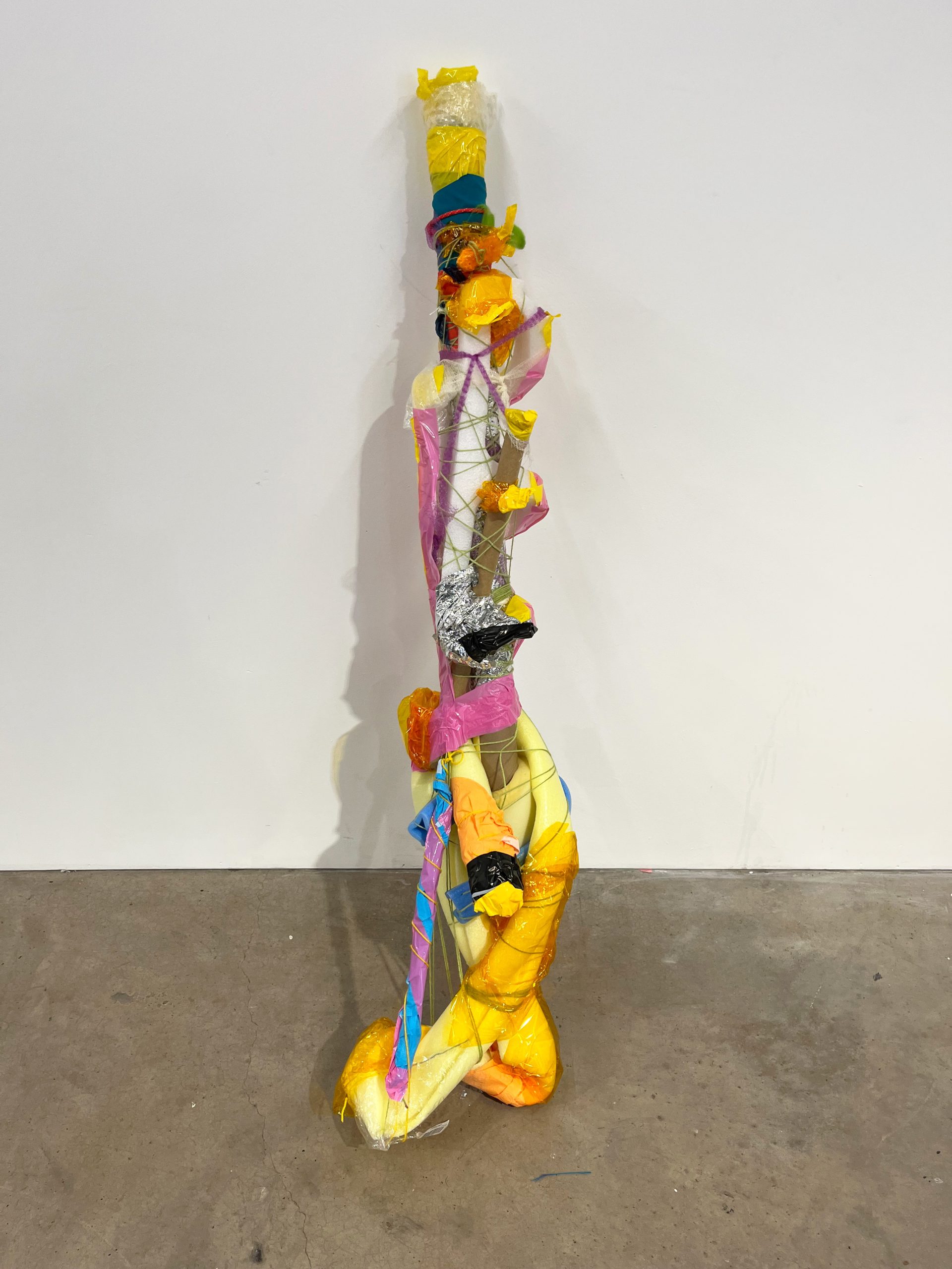 An abstract sculpture rests against a white wall on a concrete grey floor. The sculpture is assembled of various materials i.e. brown cardboard, pink and yellow tape, tinfoil. 