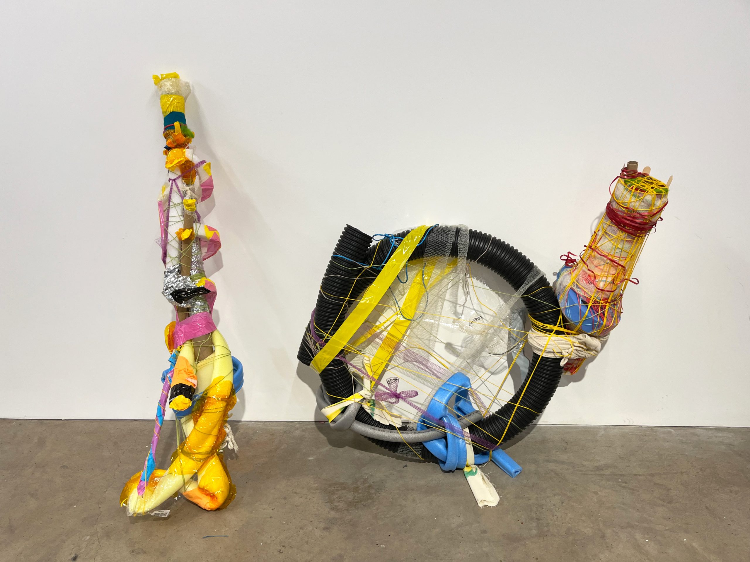 Two medium abstract sculptures rest against a white wall on a grey concrete floor. The works are assembled with colourful mixed materials.