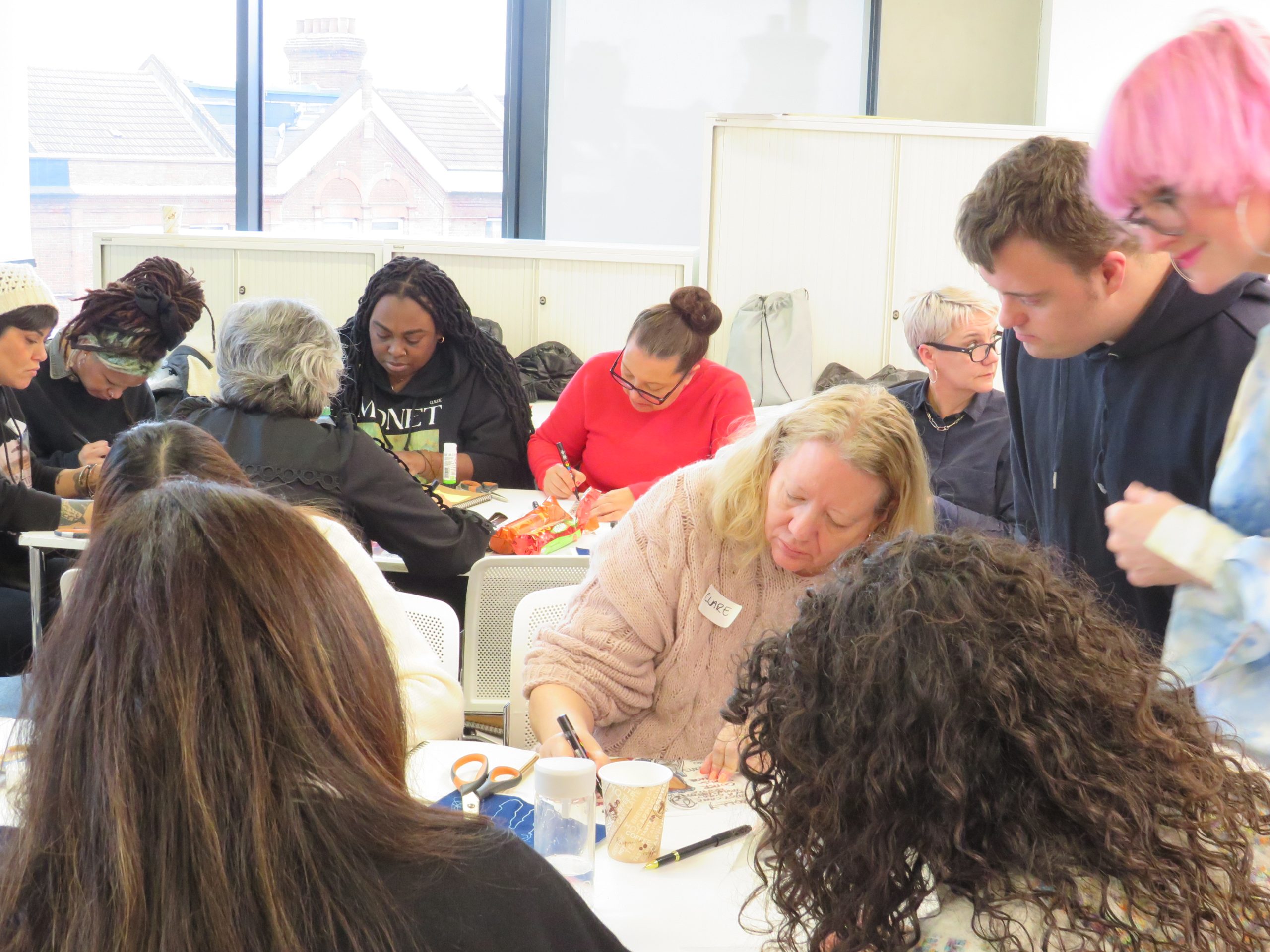 A classroom full of adult learners sat at white desks busy working. A male and female stand near guiding them in discussion.