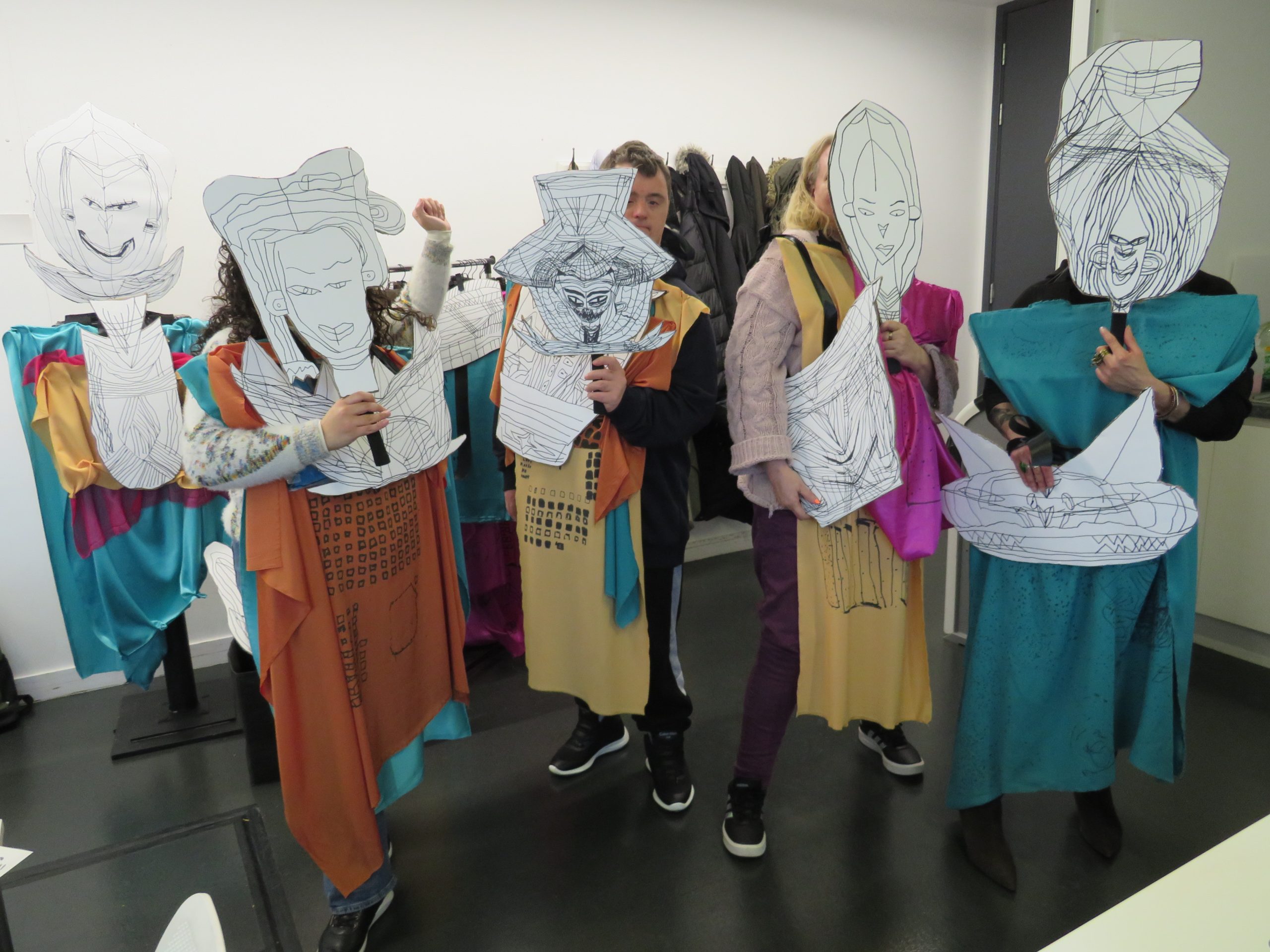 A group of people draped in colourful fabric hold pencil drawn masks in front of their faces. They are stood in a classroom.
