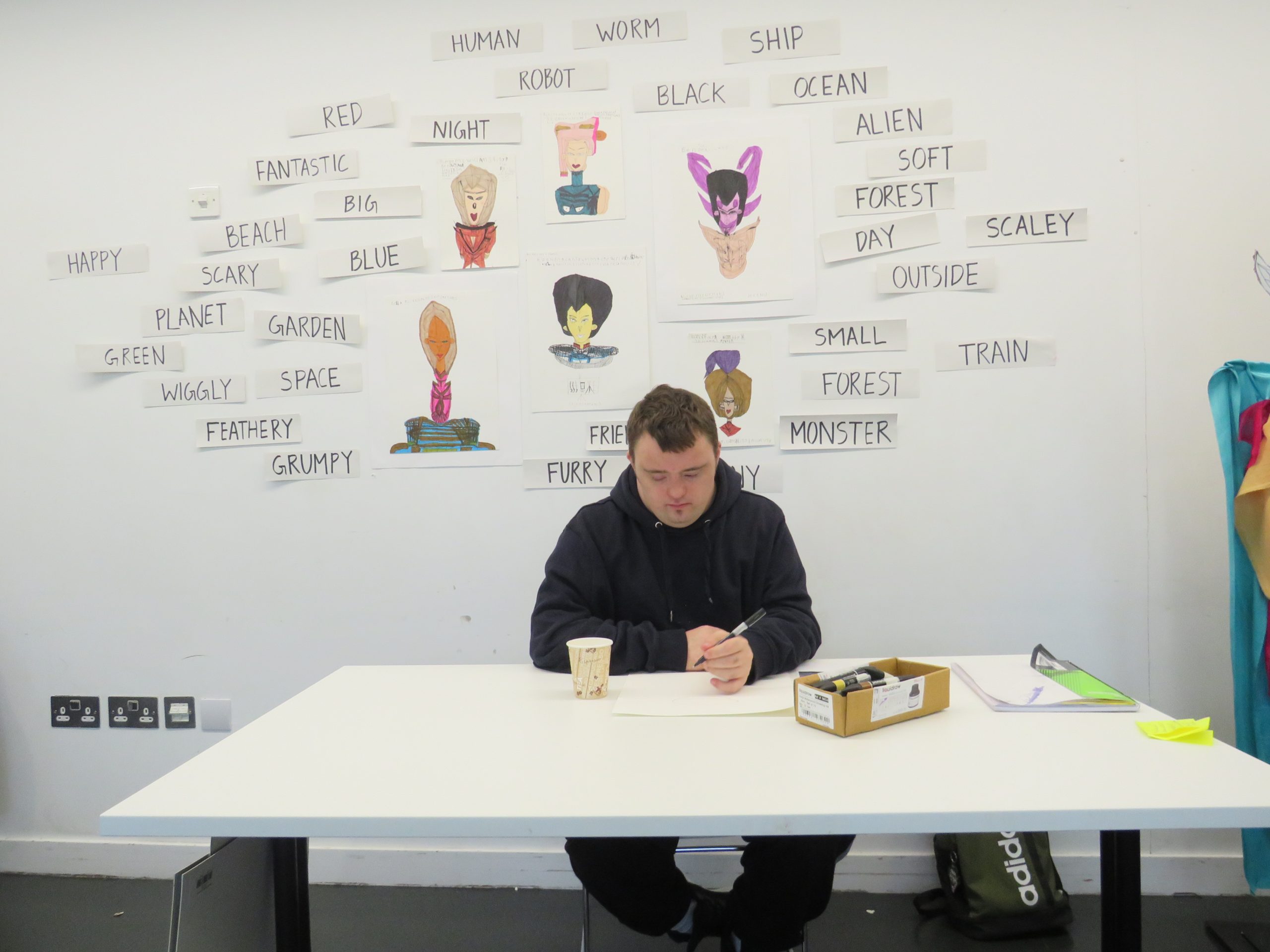 Male artist sits at a white desk working. Behind him is a white wall with graphic, fantasy drawings and text.