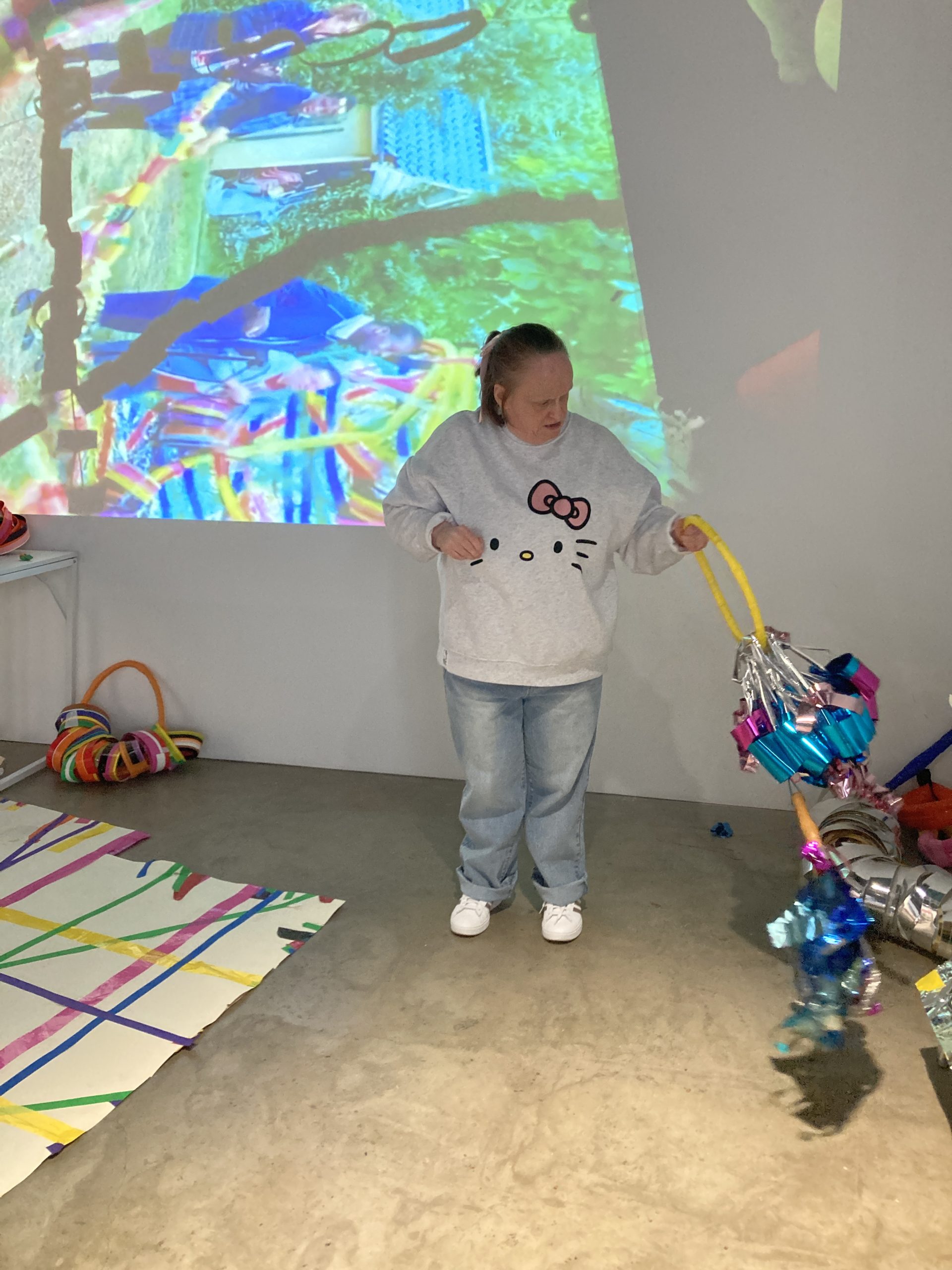 Female ActionSpace Artist Linda Bell interacts with moving sculpture. They stand in a darkly lit room with an abstract image projected onto the wall behind them. 