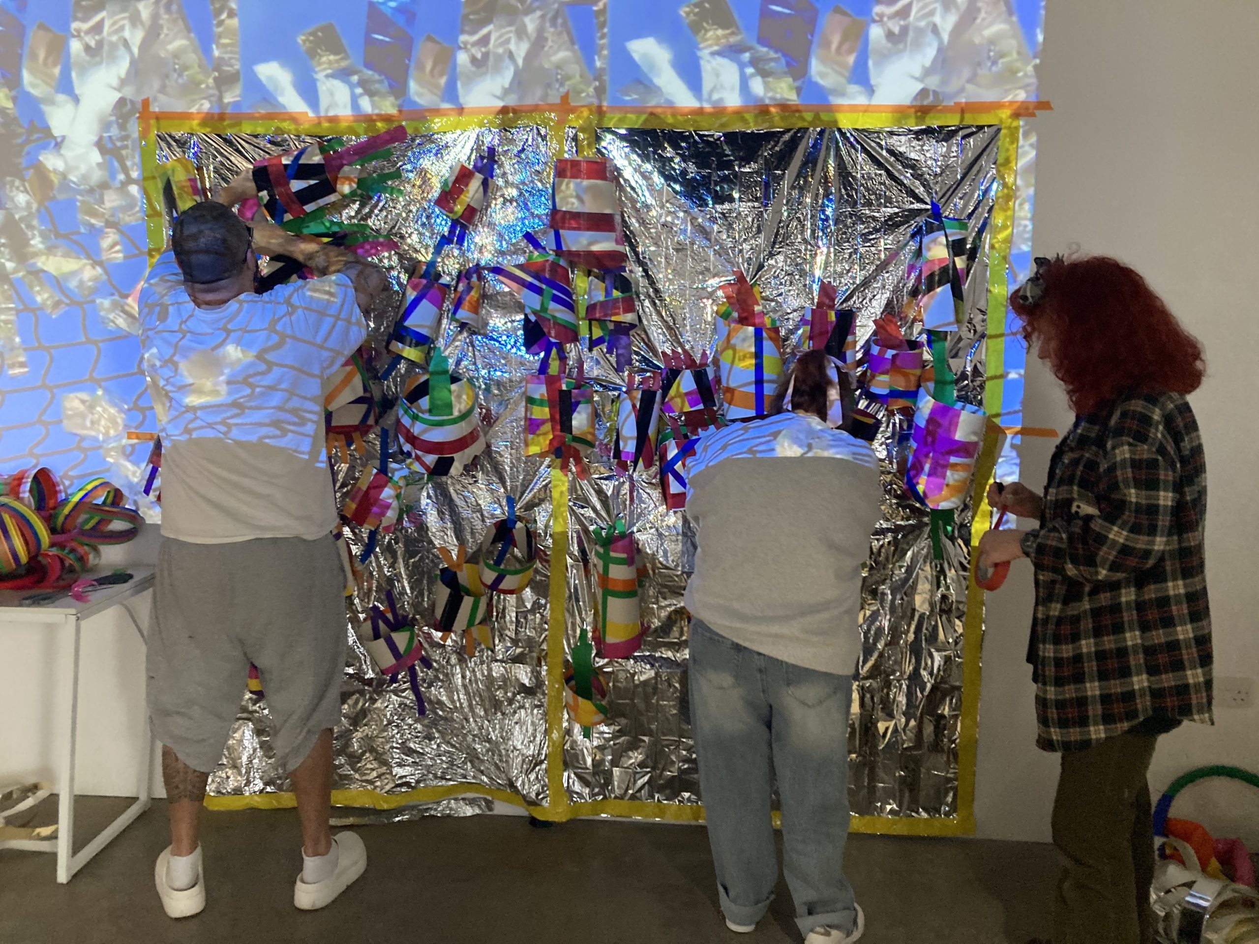 The artist, her assistant, and a participant are working on a mixed media sculpture on a wall. Their backs face the camera.