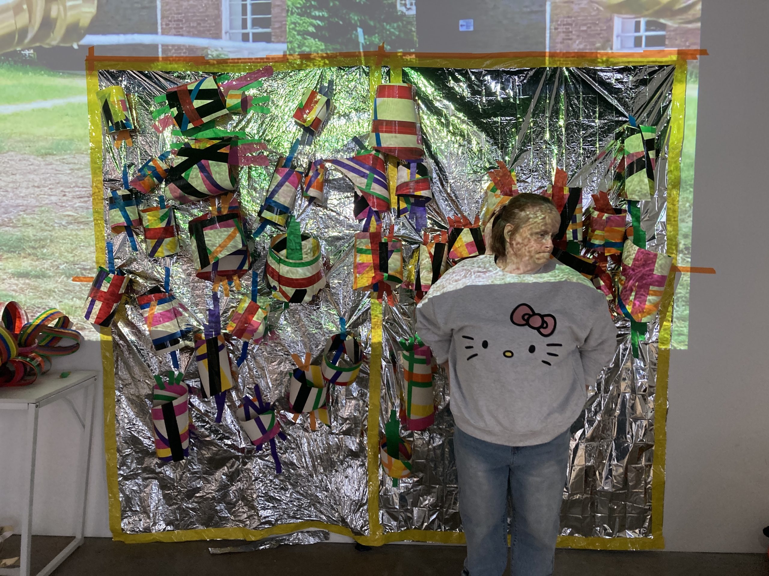 Female artist stands in front of work hung on wall in a dark room. The work is an assemblage of mixed materials with an image of a building projected across. 