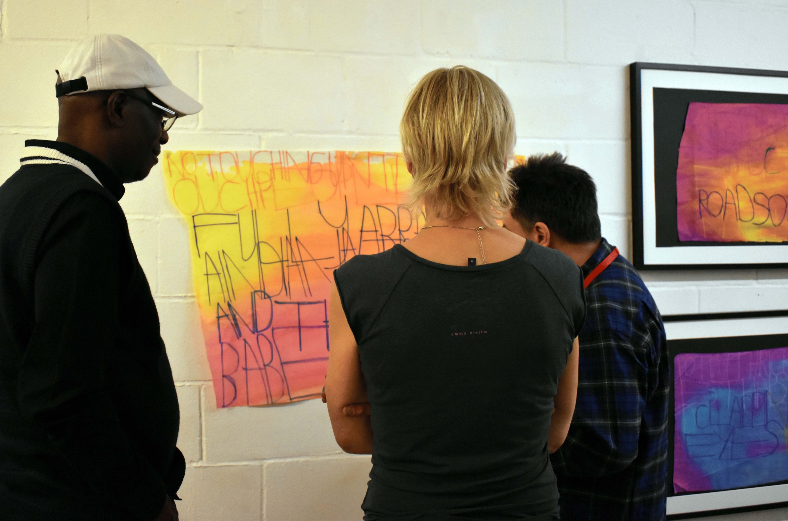 A female and two male audience members view an artwork hung on a white brick wall. Yellow and pink ink blends on the artworks fabric.