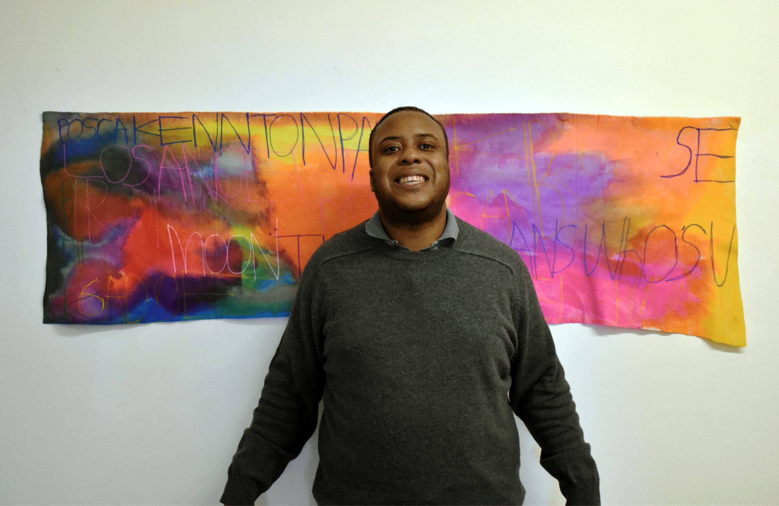 Male artist stands smiling in front of colourful artwork hung on a white wall. The work's material is fabric and ink with text written across it.