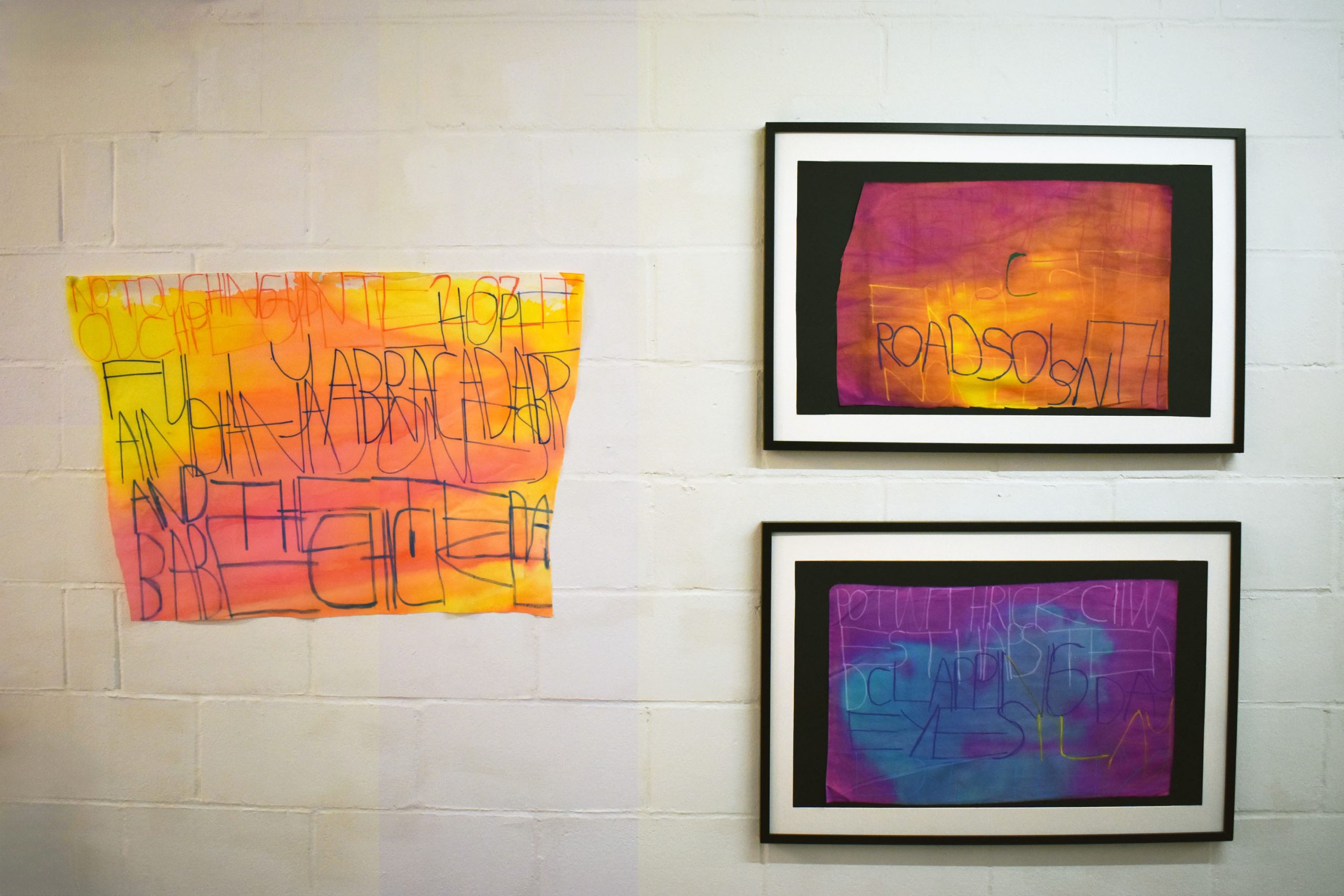 Two framed artworks and one unframed artwork hang on a white brick wall. The fabric works have colourful inks and text merging on it.