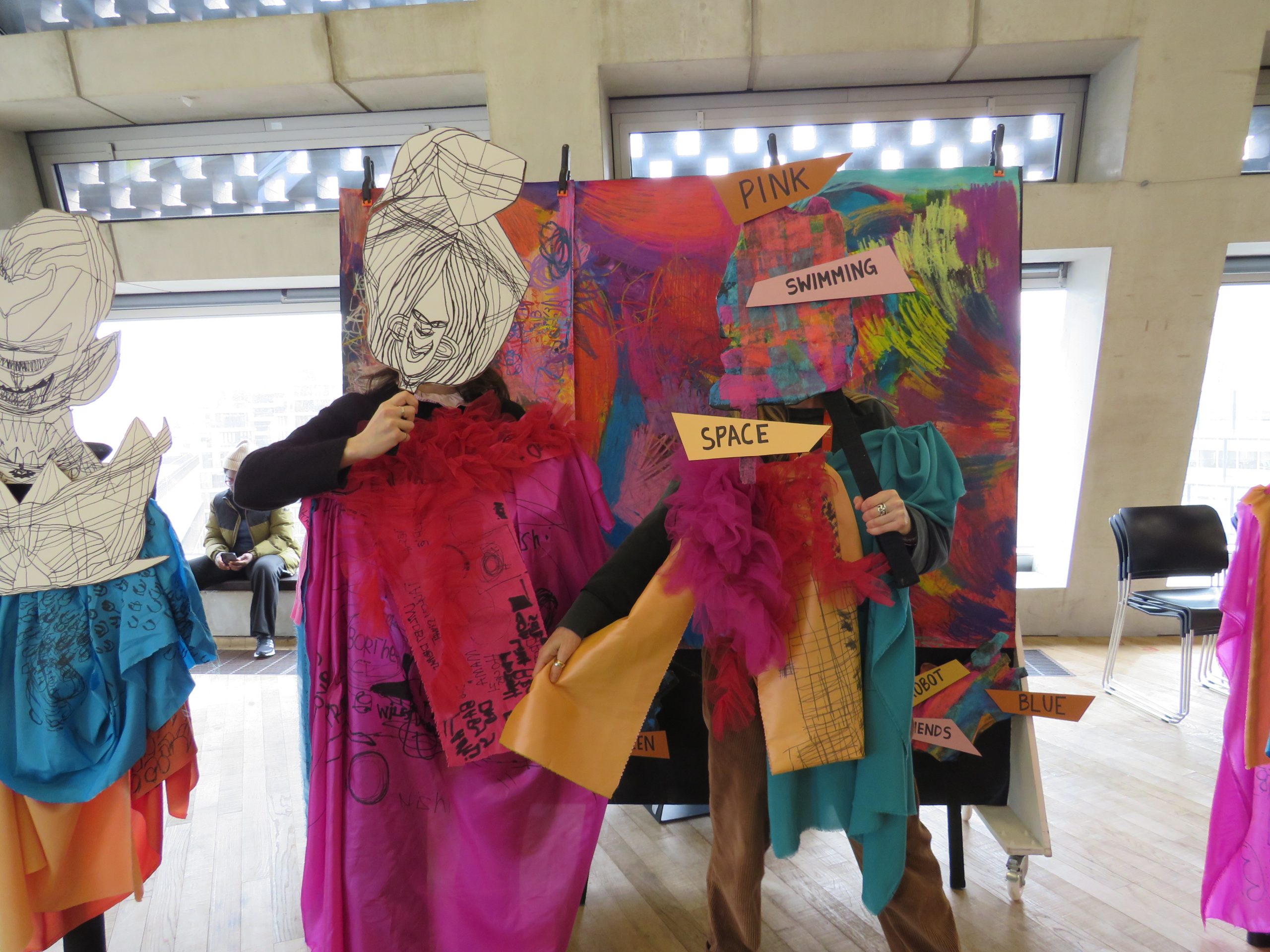 Three participants wearing costumes pose in front of a colourful backdrop in a larger room. They are holding cardboard masks in front of their faces with a array of figurative, abstract, text based drawings on.