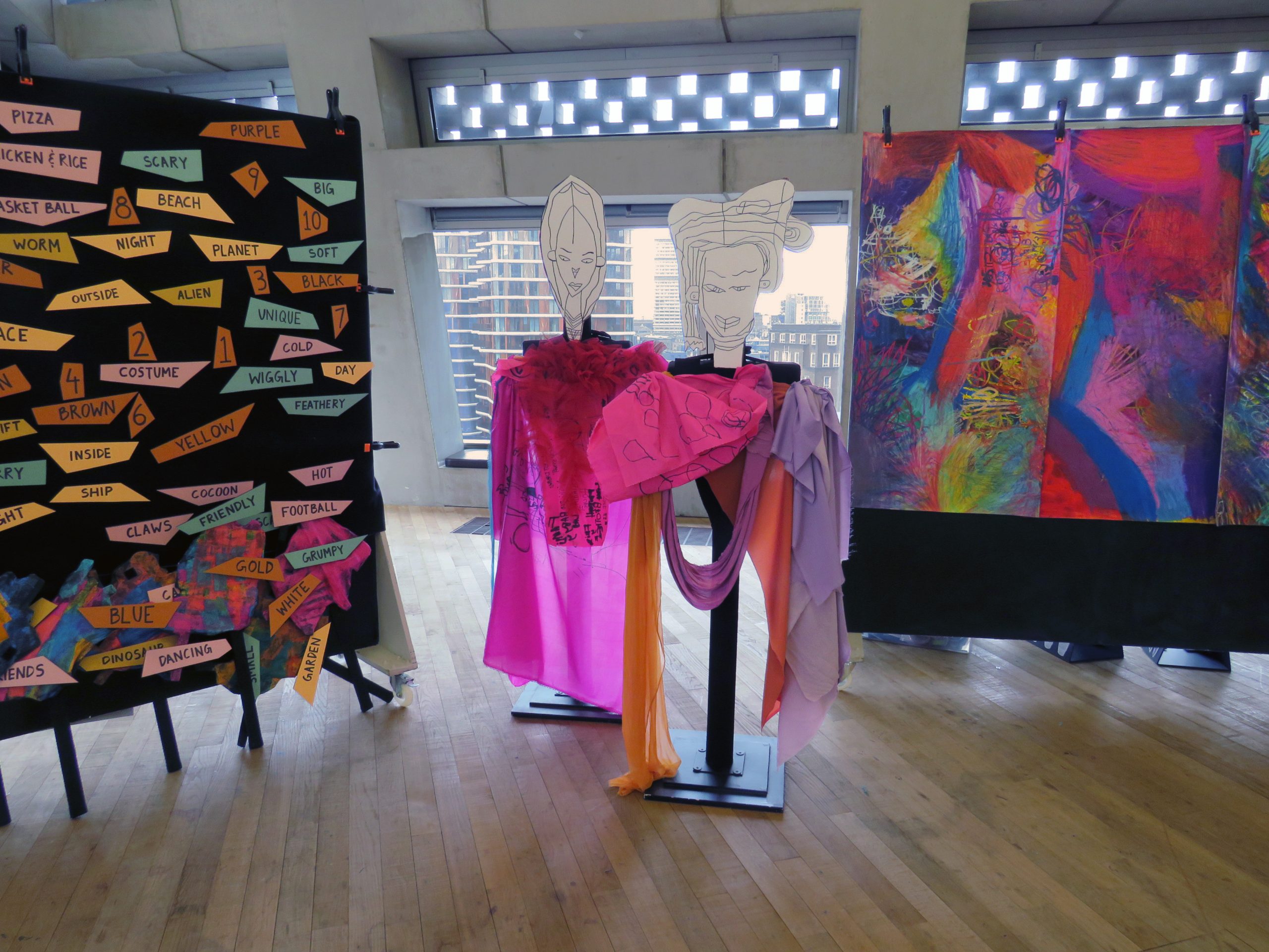 Two abstract, figurative sculptures stand in the centre of a room. On either side are display boards with colourful brushmarks and text across. 