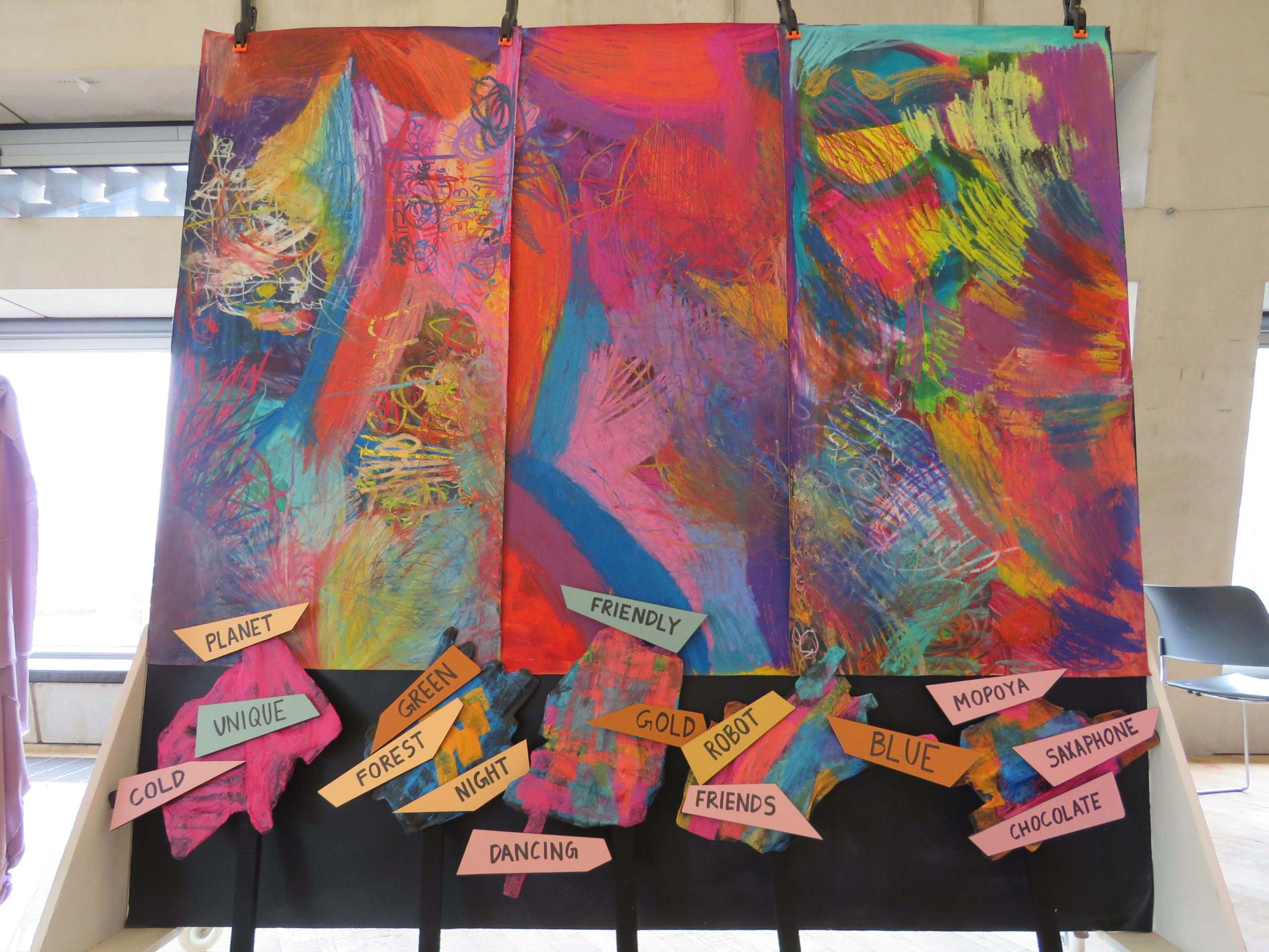 A close up of a display board with colourful blue, purple, pink, and green brushstrokes roughly painted. At the bottom the board is black with shapes stuck randomly on with text written. 