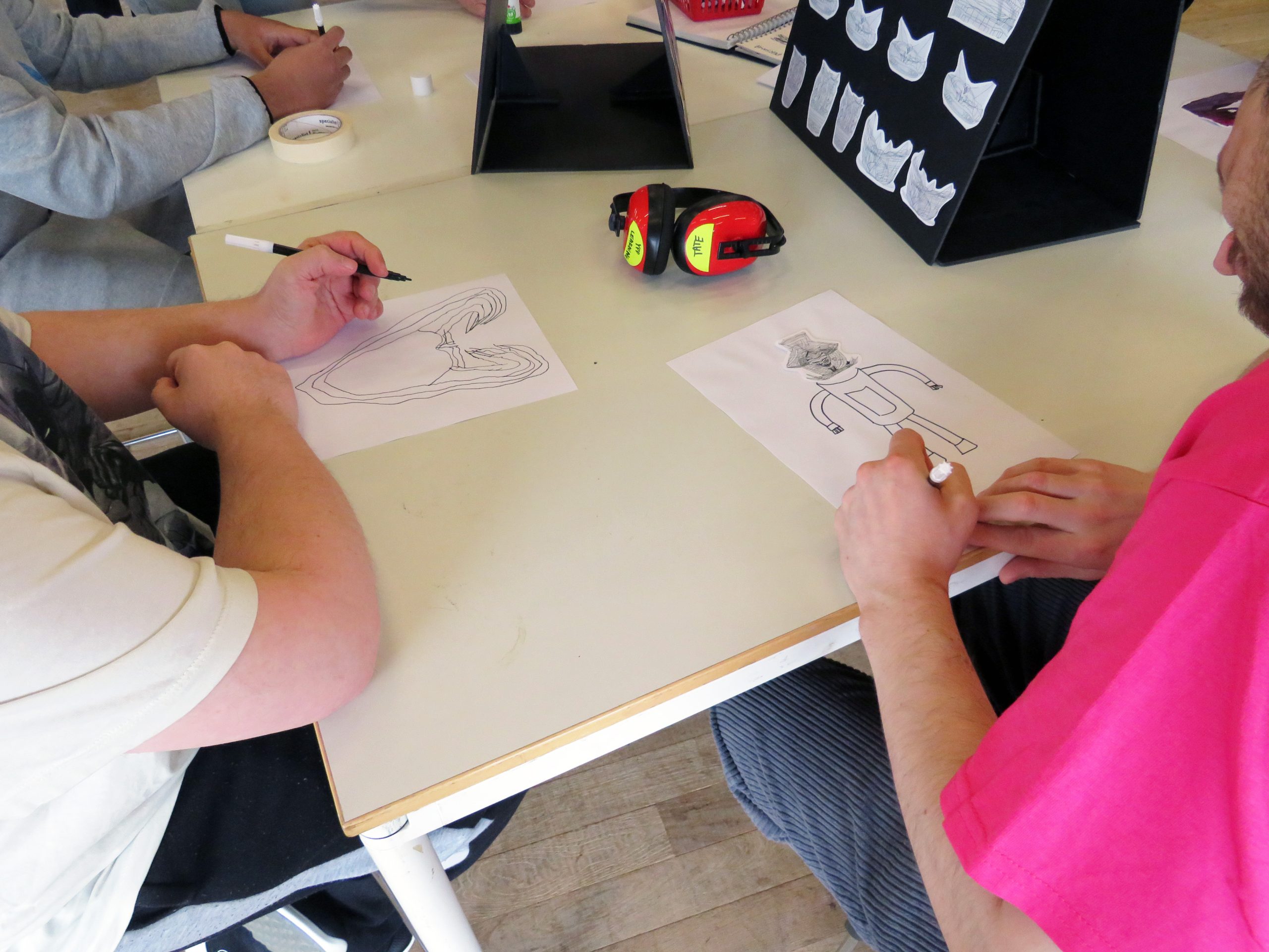 A close up of three people's arms and hands sat at a white desk. They are working on pen drawings on white paper. 