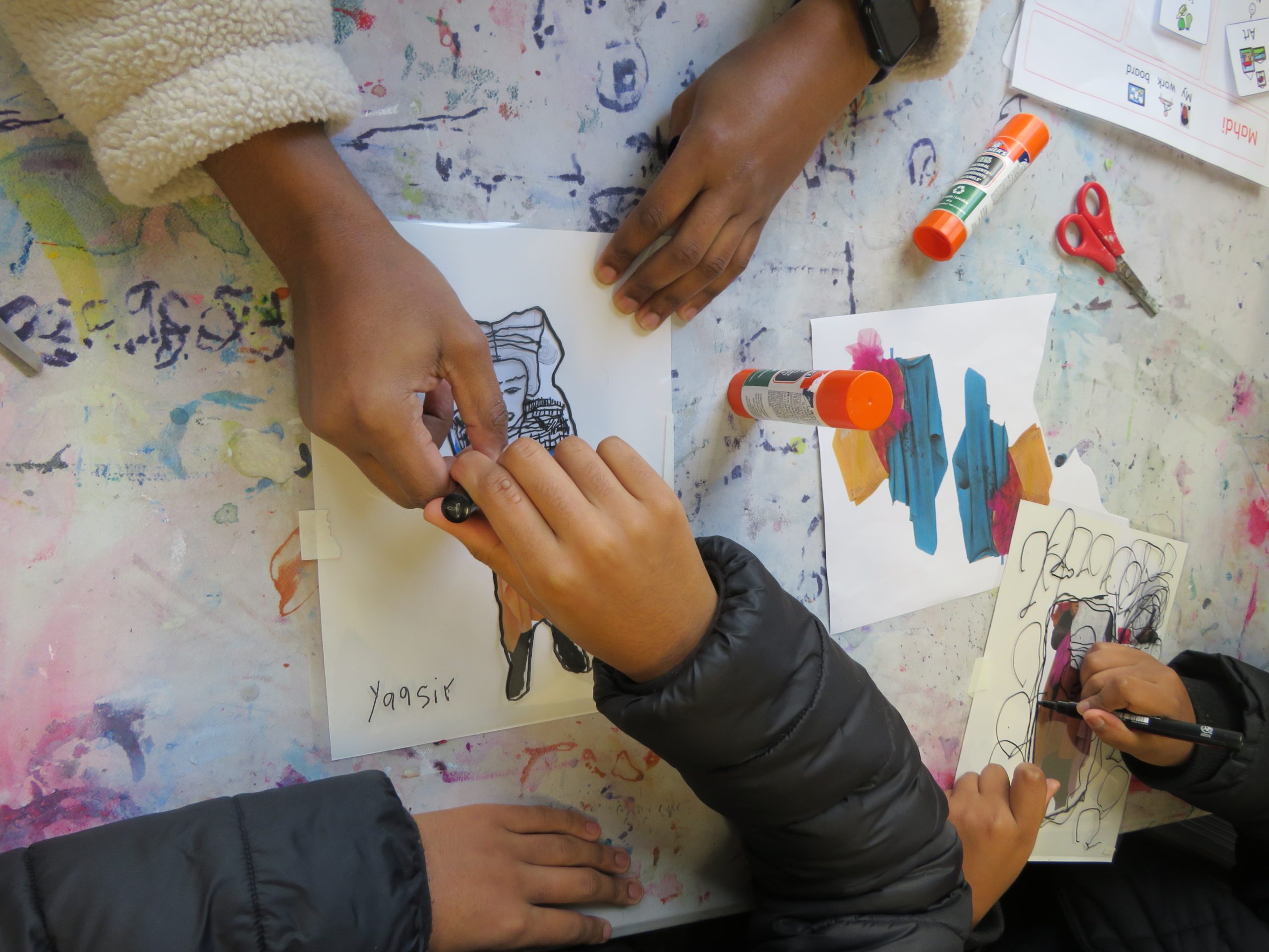 A close up of two pairs of hands working on a drawing together at a messy desk. One person passes material to another. 