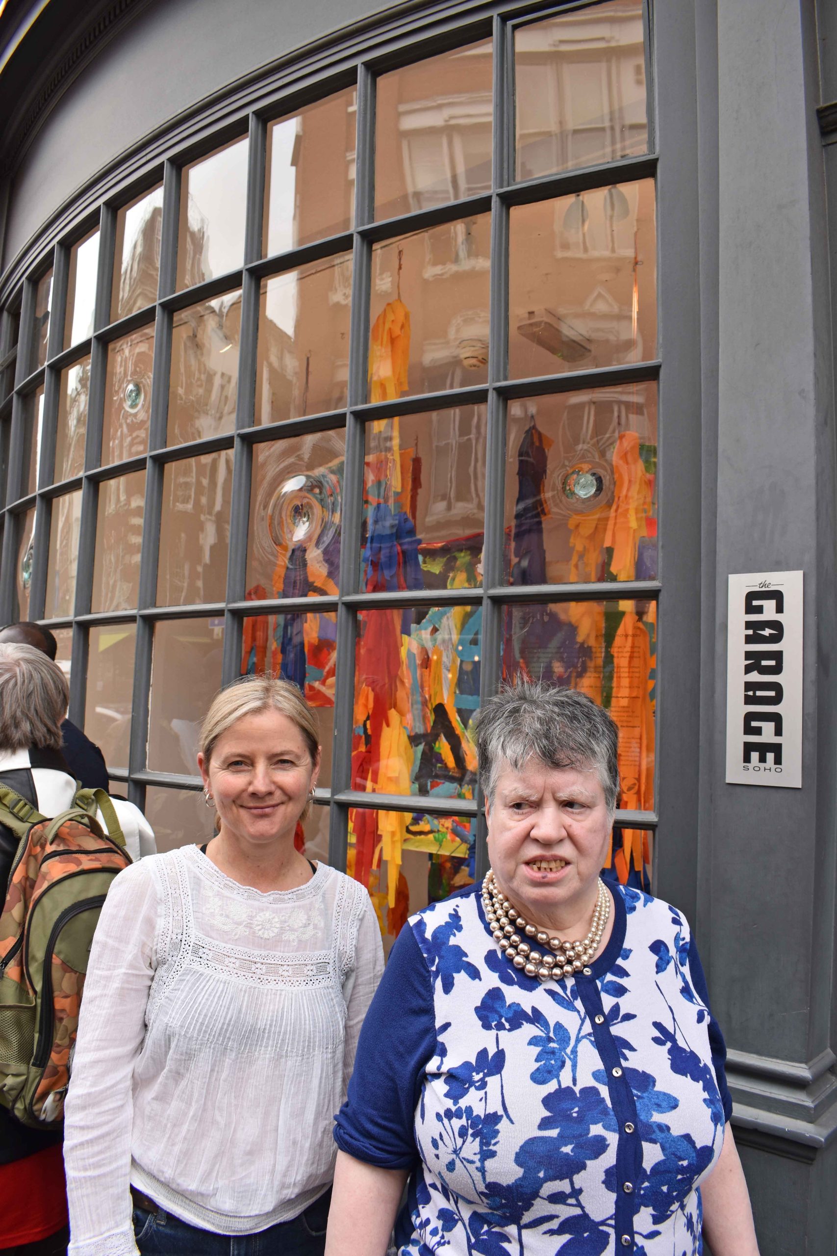 Image of two women posing for a photograph together in front of a window gallery on a street outside.