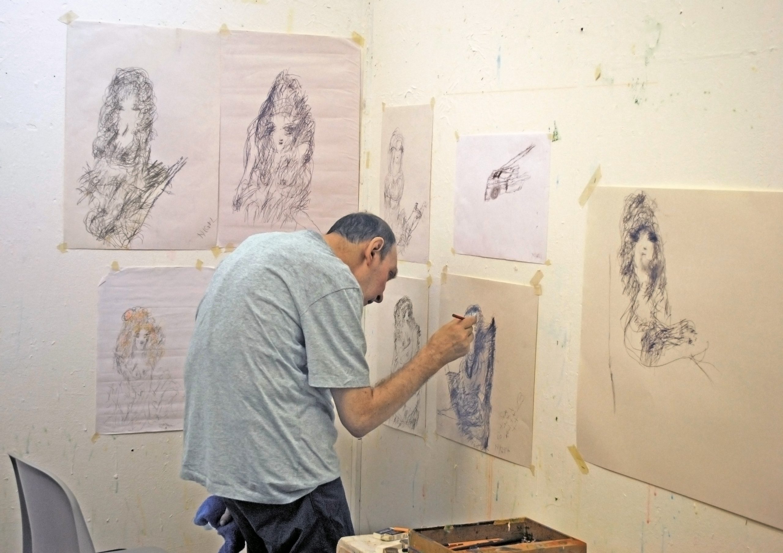 A white, male artist draws with black pencil on white paper hung against a blank white wall. Several other wispy, abstract pencil drawings are hung as well in the studio.