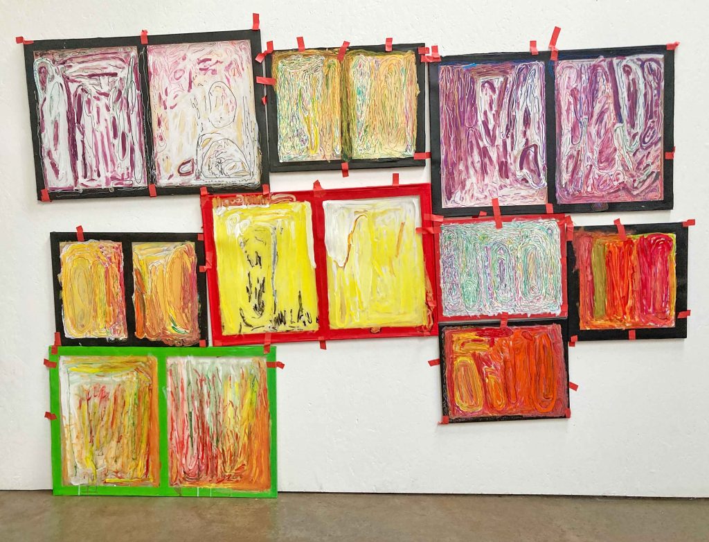 Nearly a dozen abstract paintings hang spontaneously on a blank white wall above brown flooring.