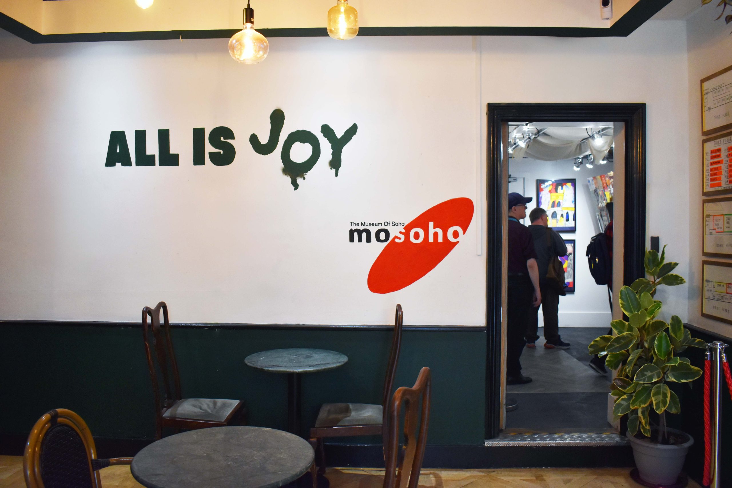  Image of an exhibition installation in a cafe/gallery space. On the wall is a sign reading 'All is Joy' and 'Museum of Soho'.
