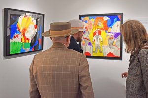Image of three people looking at framed artworks hung on white walls. The works are graphic, coloured pencil drawings with political references.