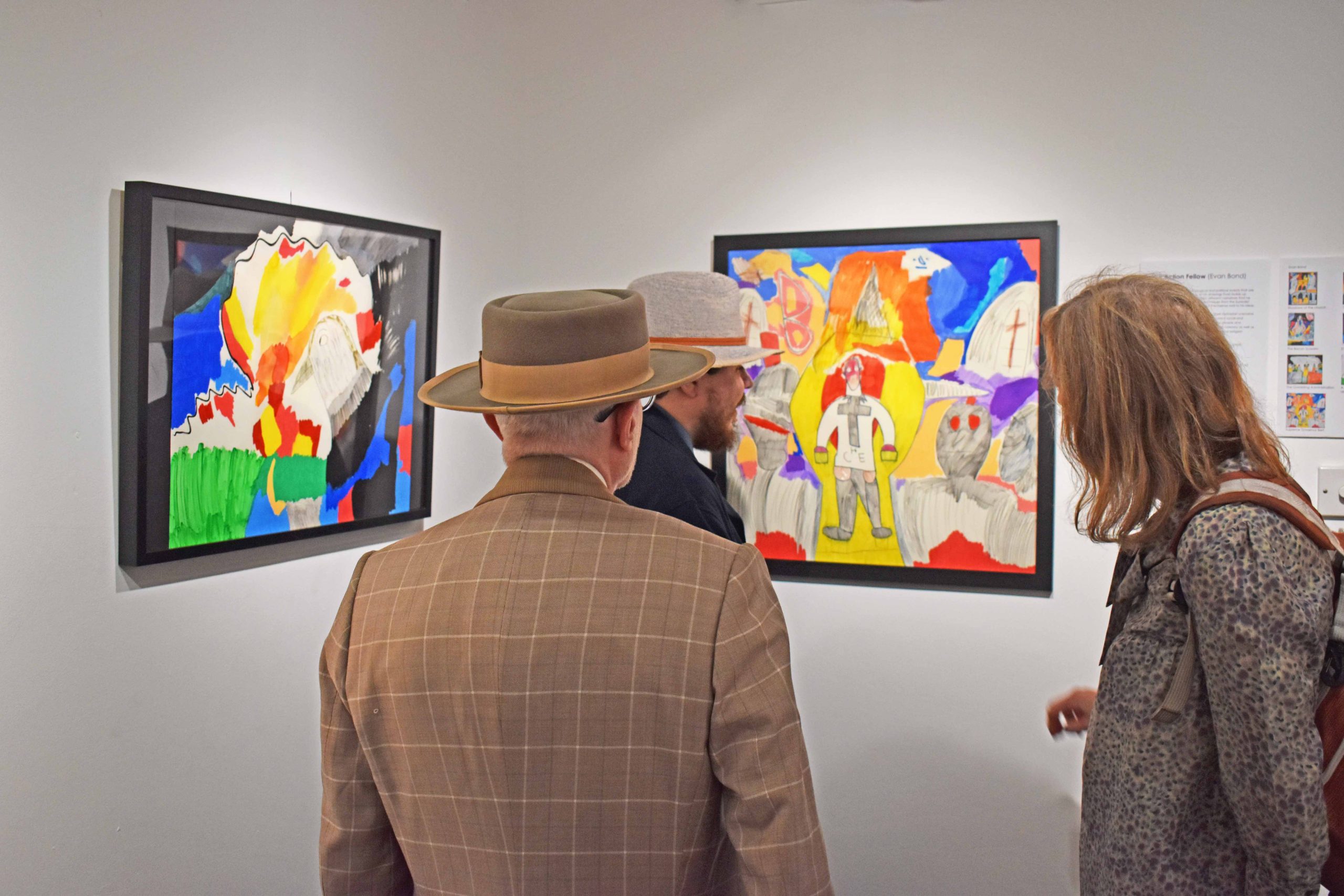 Image of three people looking at framed artworks hung on white walls. The works are graphic, coloured pencil drawings with political references.