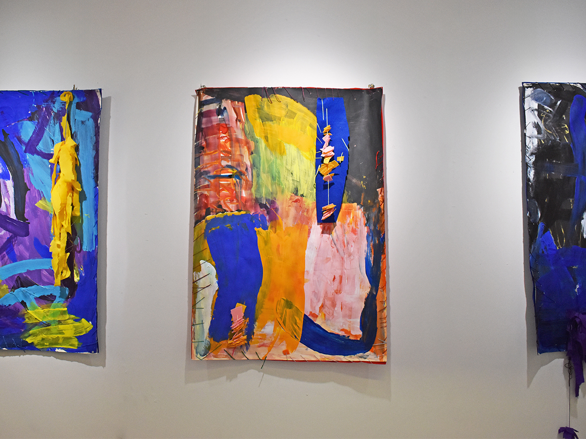 Image of three mixed media artworks hung on a white wall. The artworks are medium, portrait on paper with paint, drawing, and textiles incorporated.