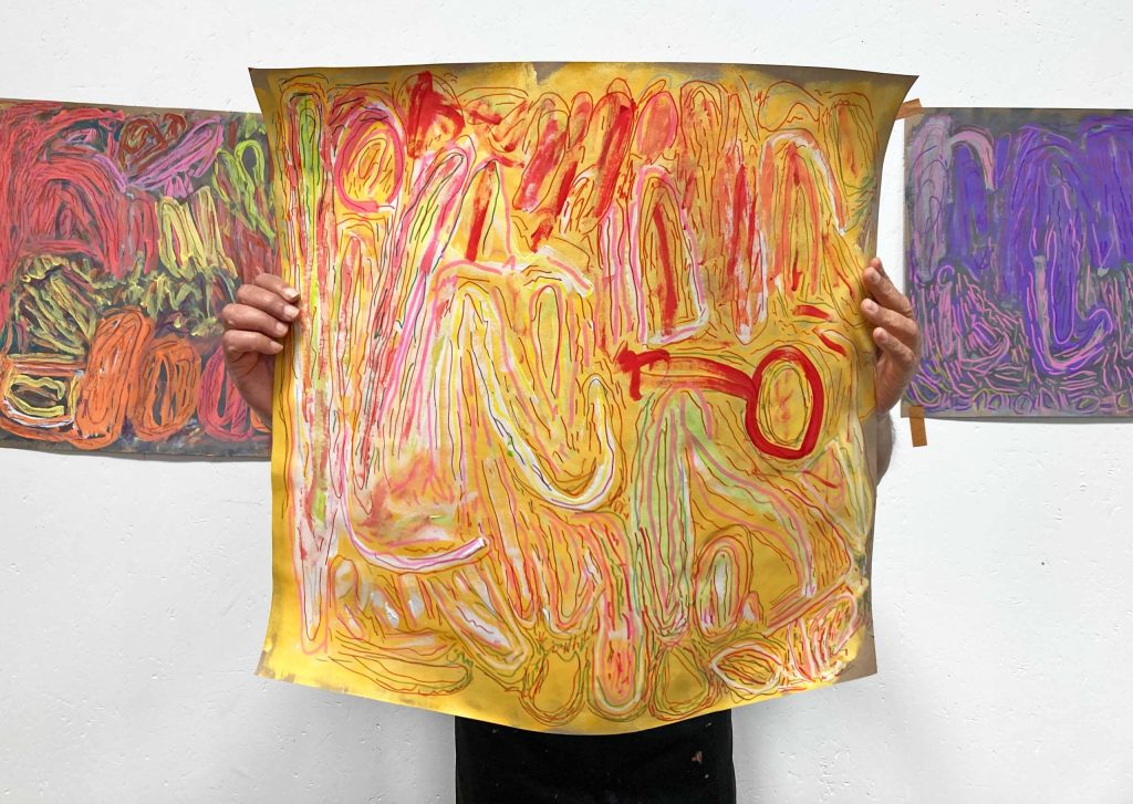 Artist holds artwork in front of them, covering their face and upper body. Abstract paintings hang on a blank white wall behind.