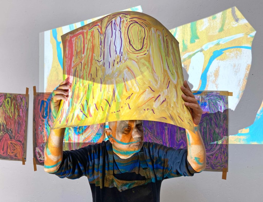 In a darkly lit room an artist holds artwork in front of them, covering their face. Abstract paintings hang behind and projected art floods the scene.