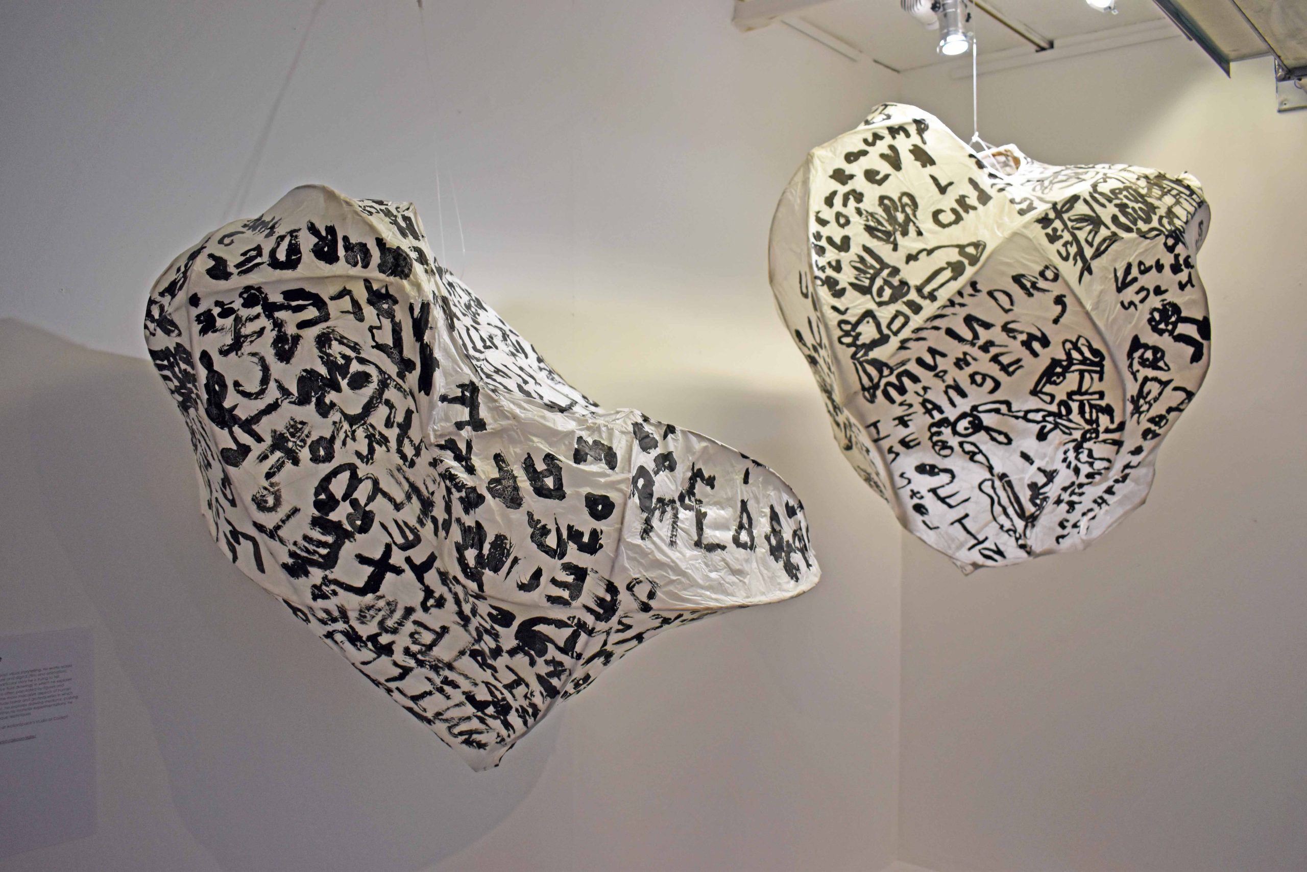 Two monochrome sculptures hang mid air, suspended from the ceiling in a white gallery space. The abstract sculptures have black text and lettering painted on. 