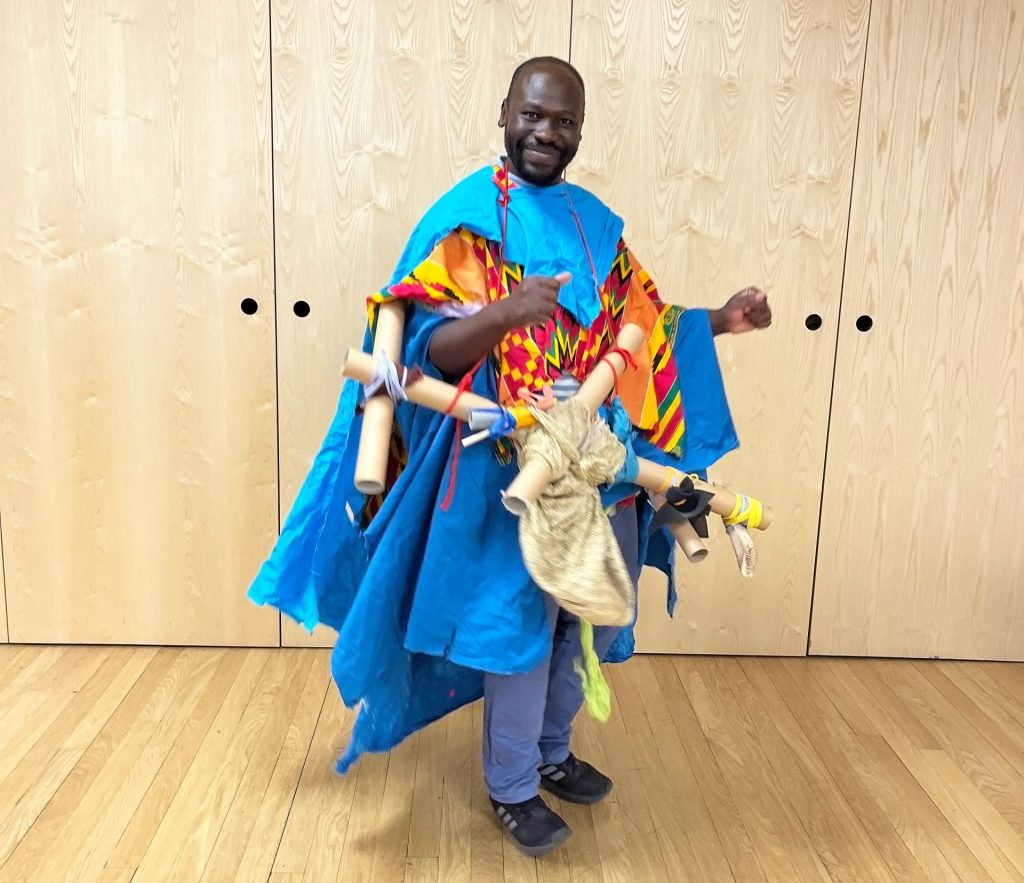 Black male artist wearing vibrant, colourful costume holds interactive cardboard sculpture.