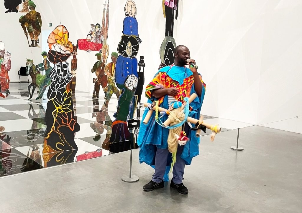 Black male artist in colourful costume holds cardboard sculpture in a gallery space.