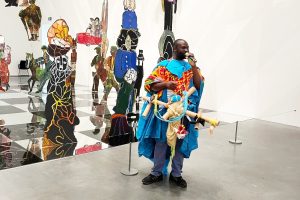Black male artist in colourful costume holds cardboard sculpture in a gallery space.