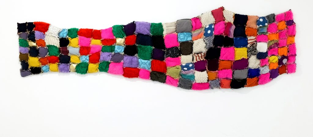 A textile artwork hanging horizontally on a blank white wall. Dozens of colourful textile patches of varying fabrics are sewn together in a soft, grid-like fashion.