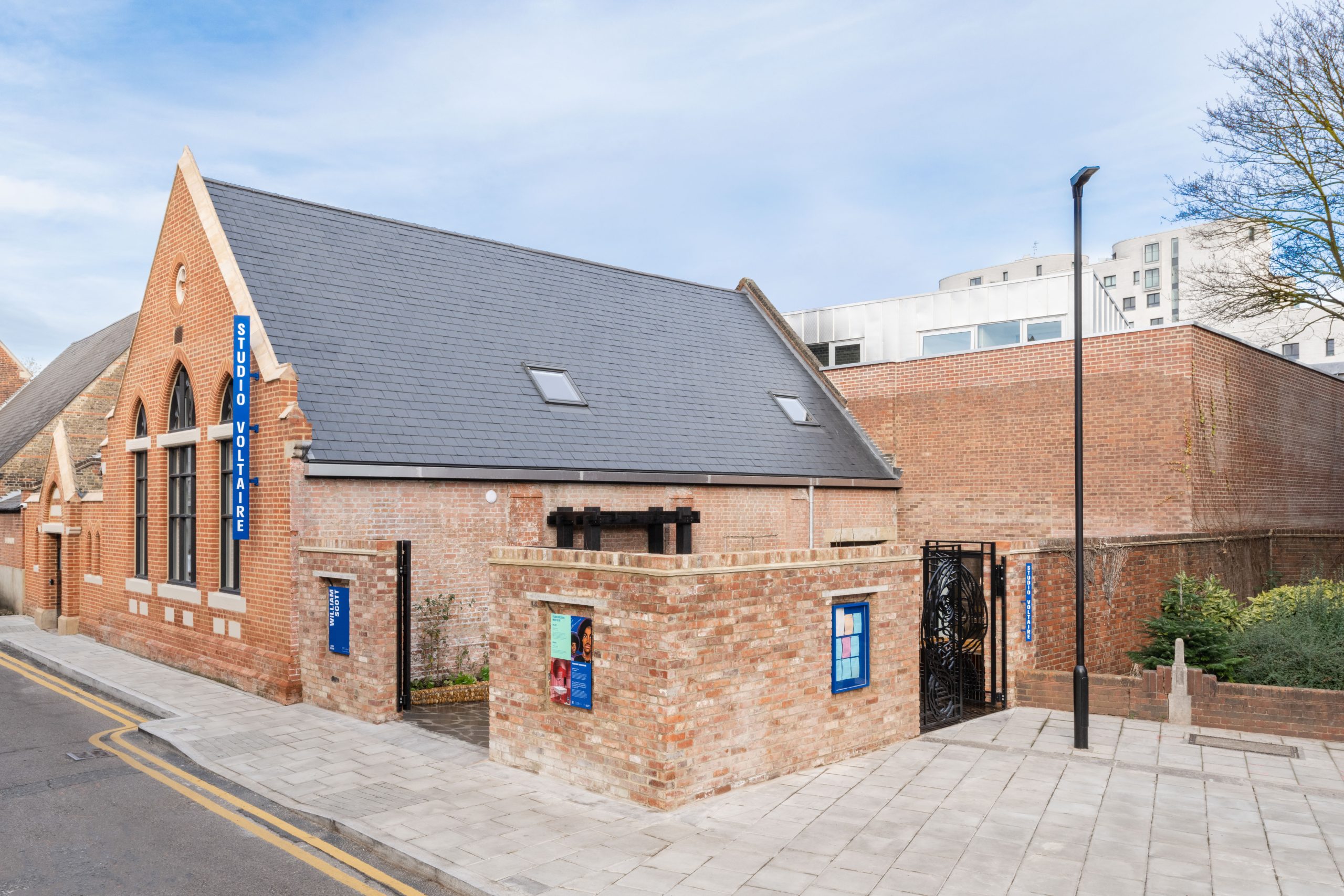 Street view of the entrance to renovated church hall building with orange brick, black wrought iron gating, and colourful blue signage outside.
