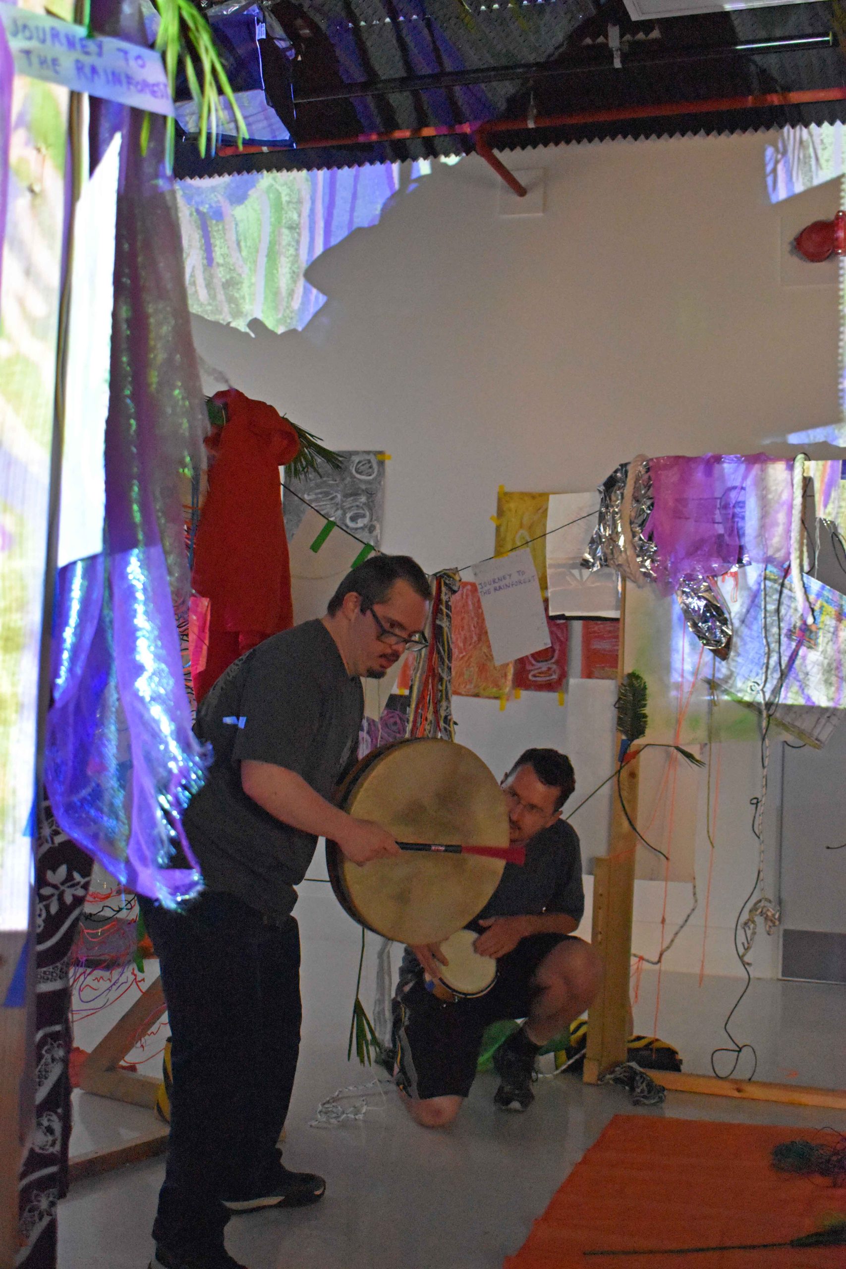 ActionSpace artist James Heath hits a drum looking off camera with Facilitator Louis Caseley.
