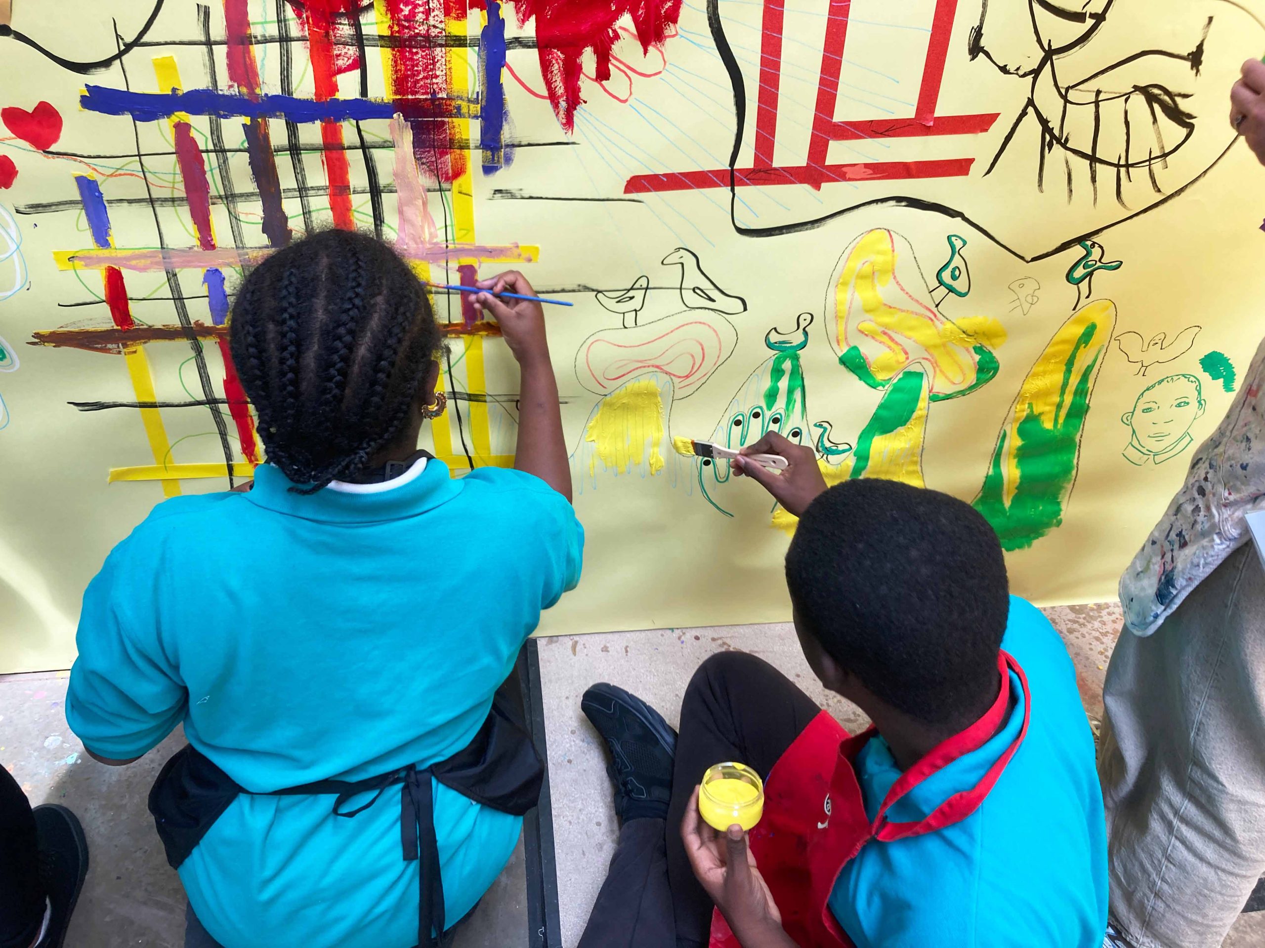 A young black girl and boy in uniform sit on the floor painting art on a yellow wall