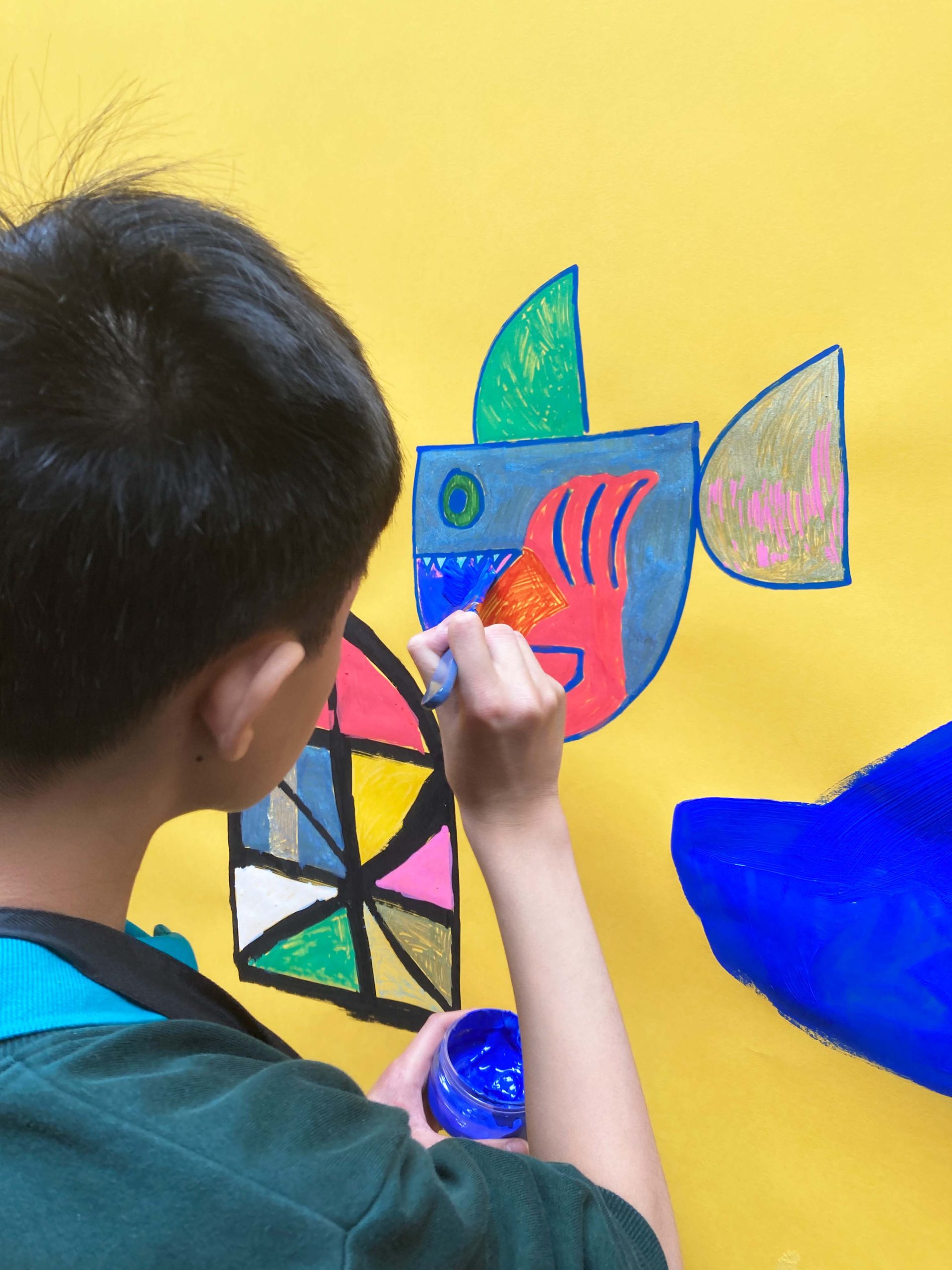 The back of a young white boy's head and he draws a colourful, asbtract fish in pen on a yellow wall.