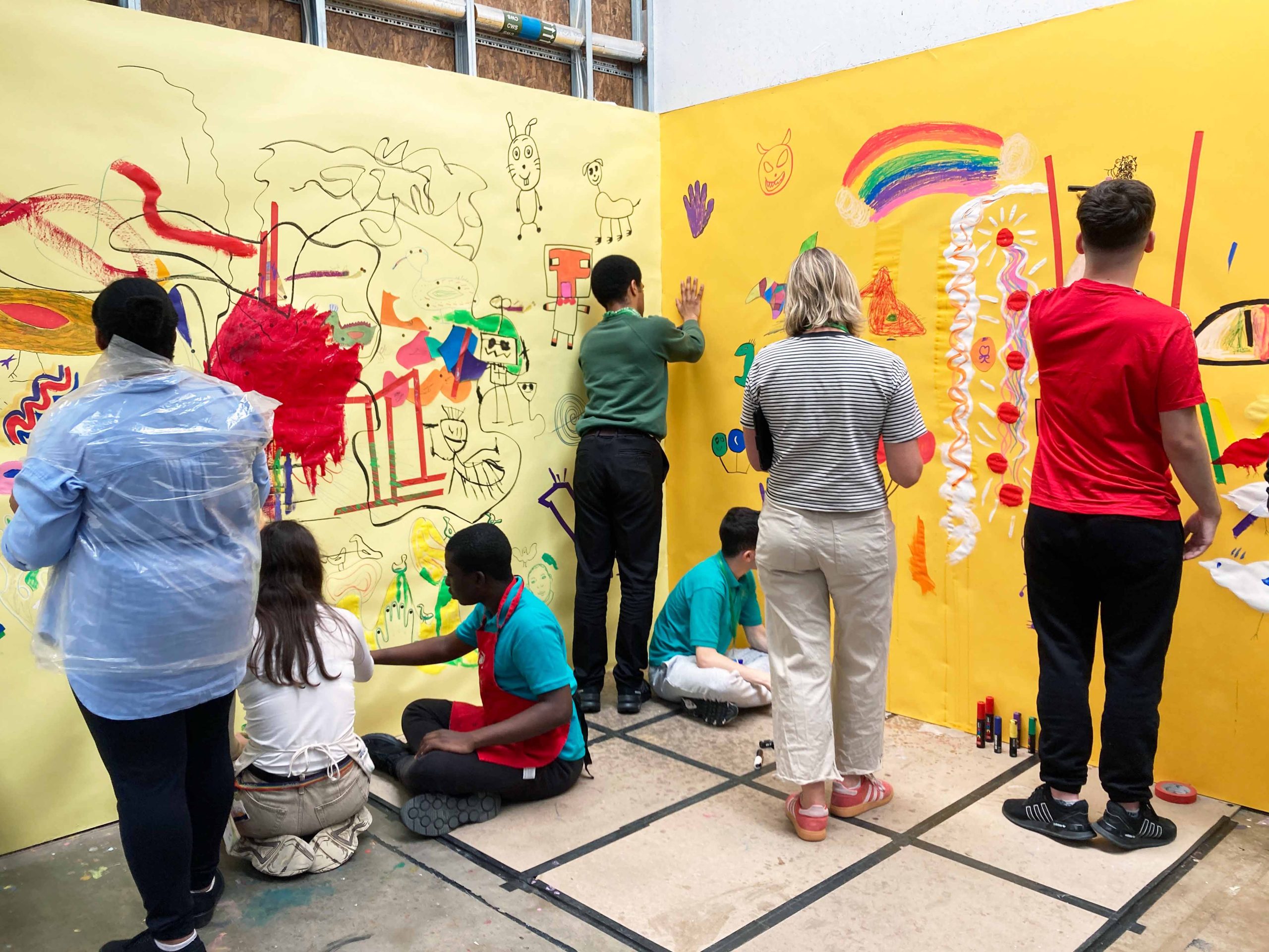 A group of children and one female adult face a yellow wall drawing colourful pictures onto it.