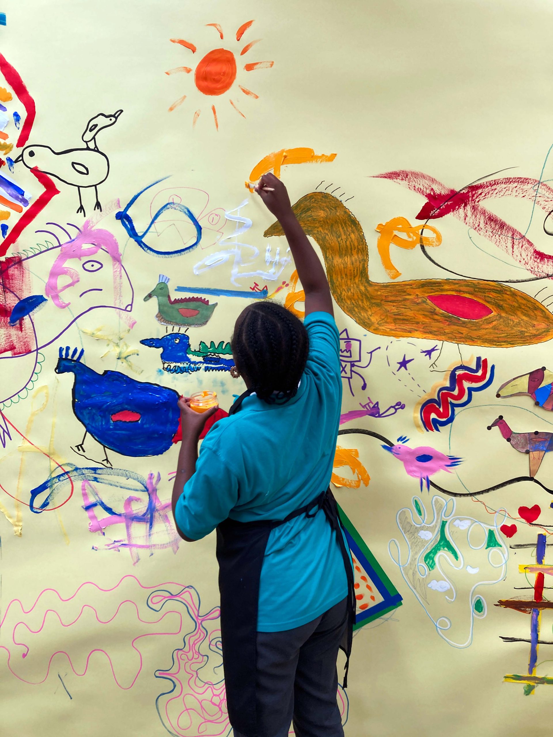 A young black girl wearing uniform reaches up to make marks on a wall covered in drawings.