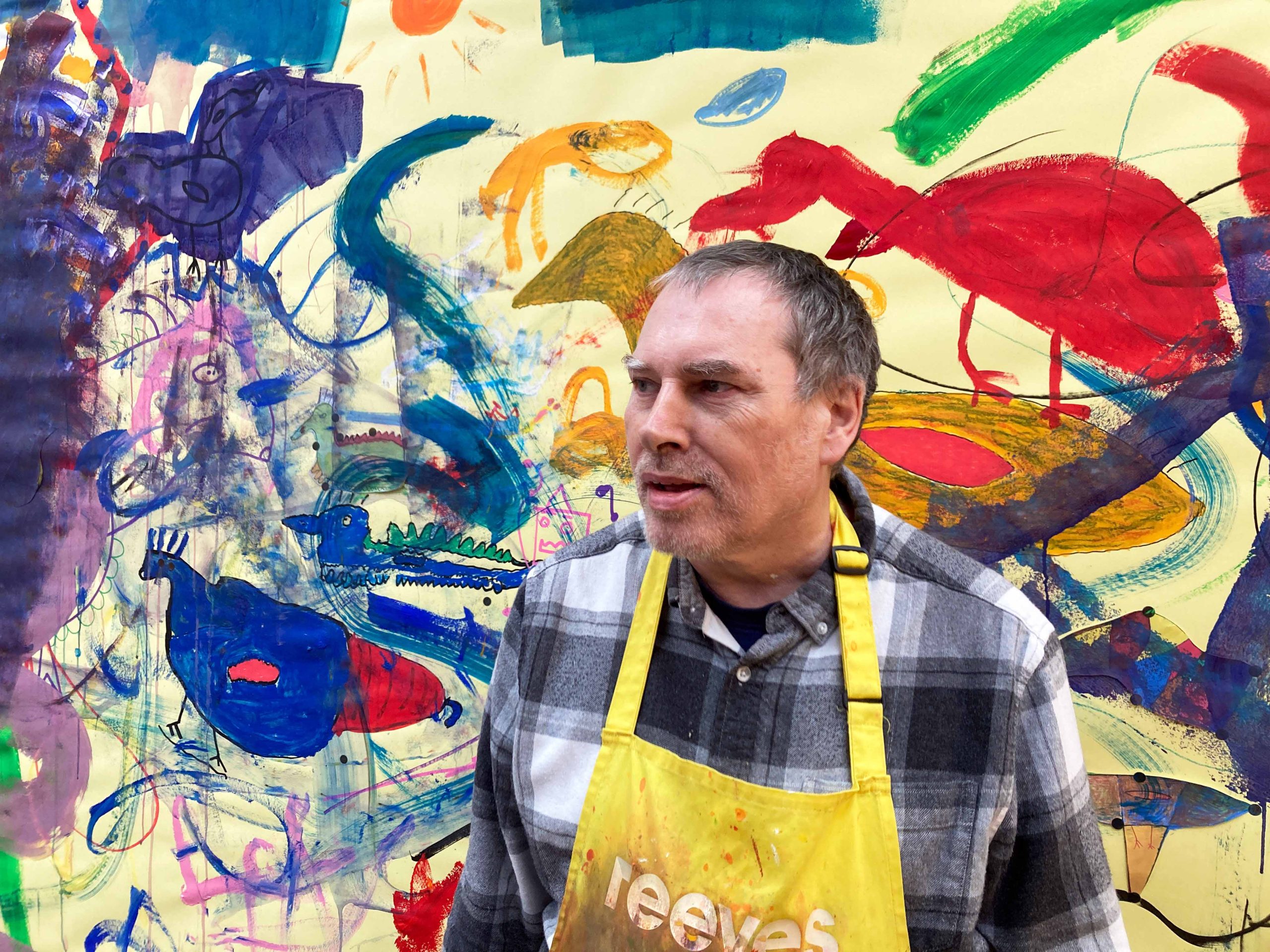 Image of artist Robin Smith looking to the left of the camera with a vibrant yellow wall behind him with figures and shapes painted on top.