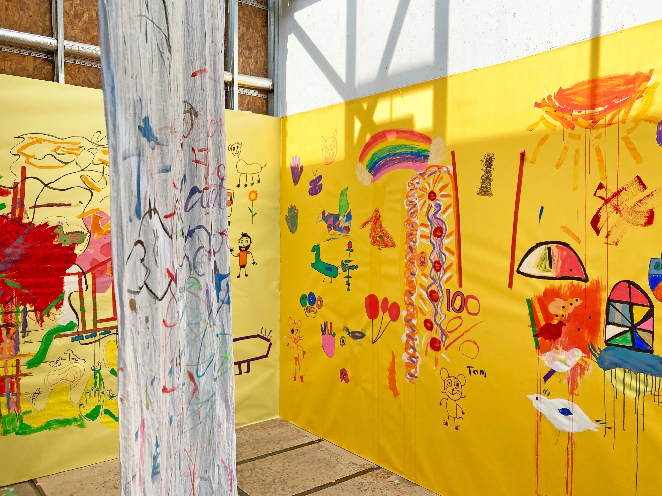 A large white cloth covered in colourful drawings hangs from a ceiling in a room with yellow walls and various drawings on it.