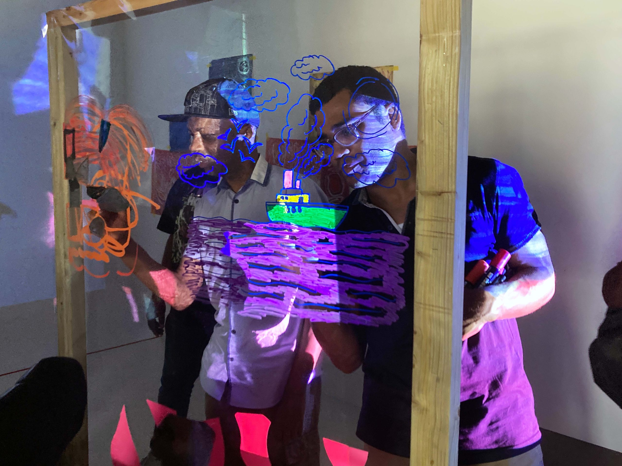 Audience members draw onto see through material on a freestanding wooden frame in a dark space.