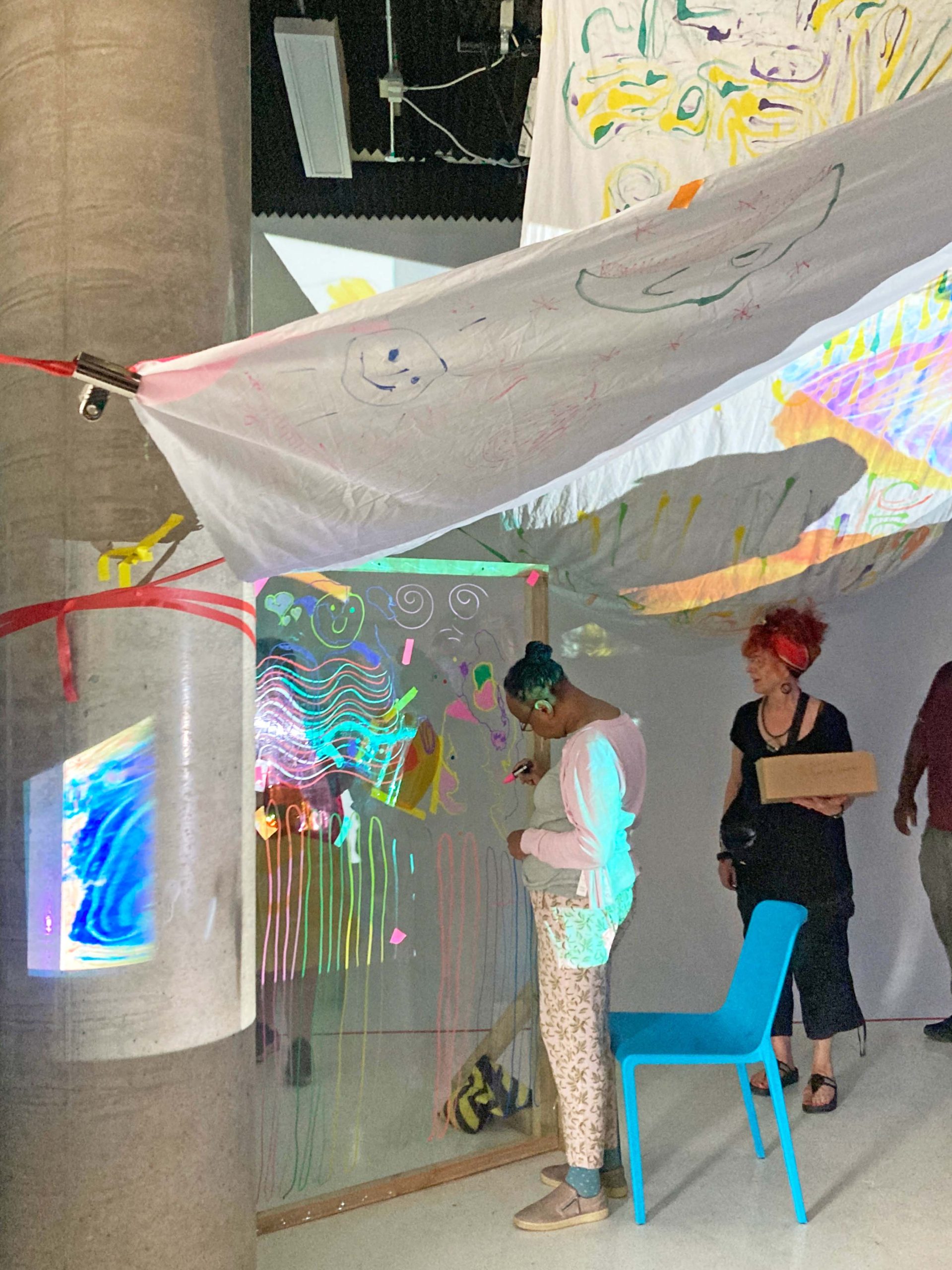 Audience members watch as others draw onto large see through material in a art space.