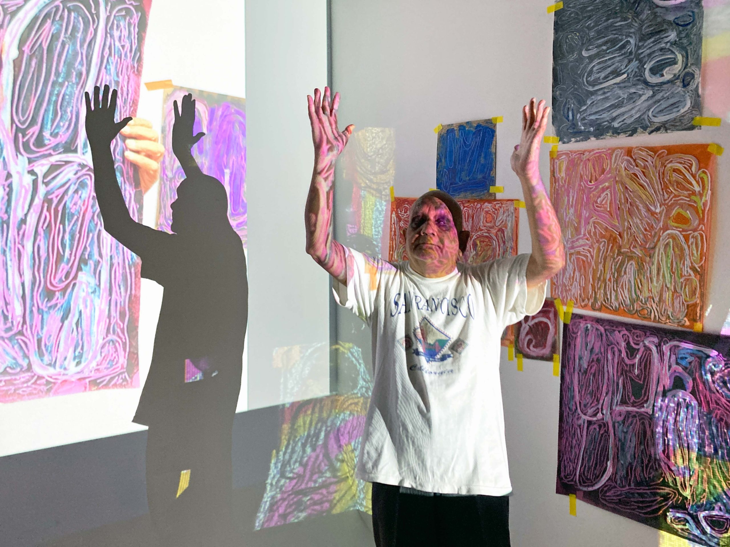 ActionSpace artist Chandrakant Patel holds his hands up to the sky with projected artworks on the wall behind.