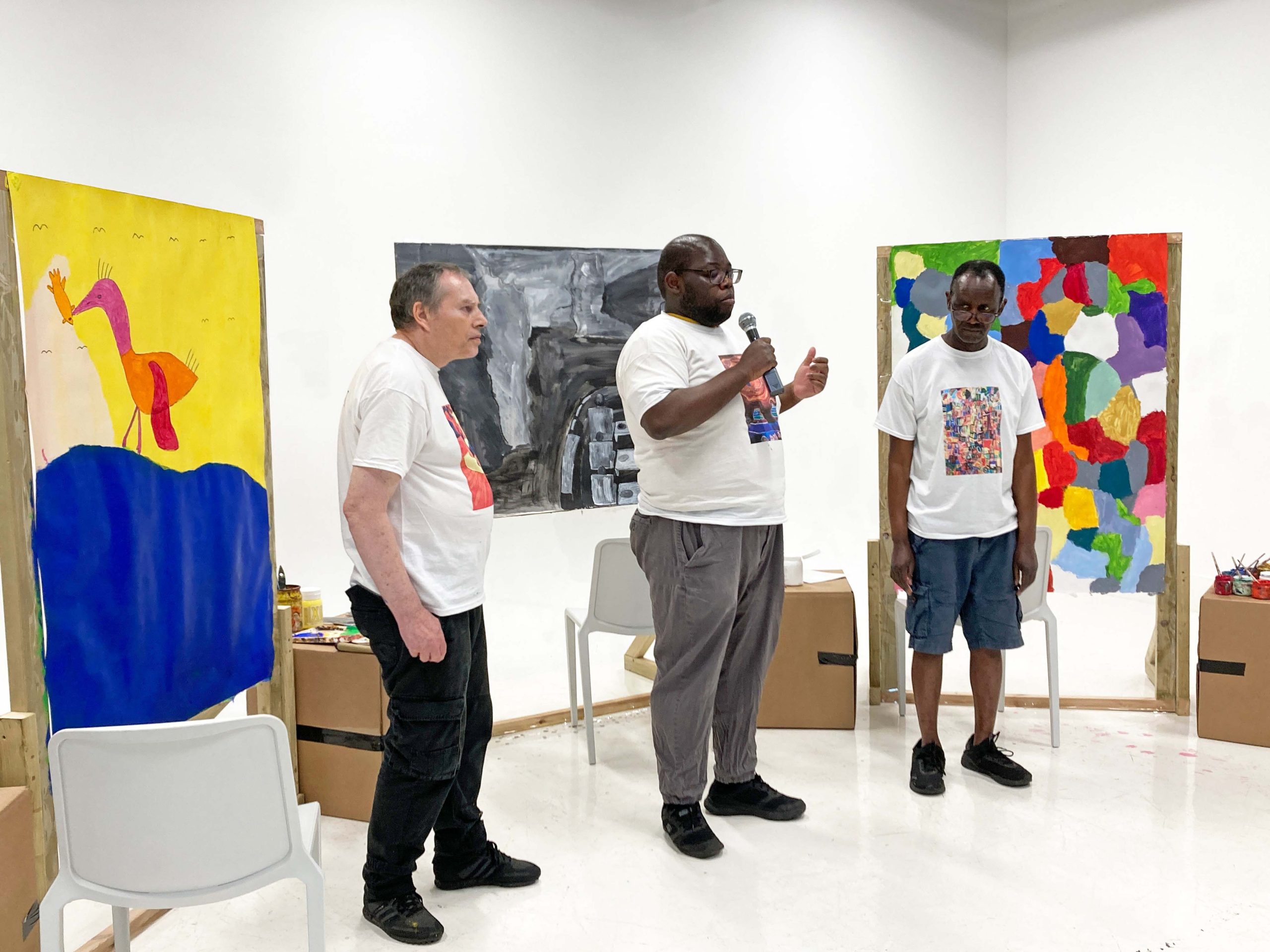 Three male painters stand in front of their works, one speaks into a microphone to an audience off camera.
